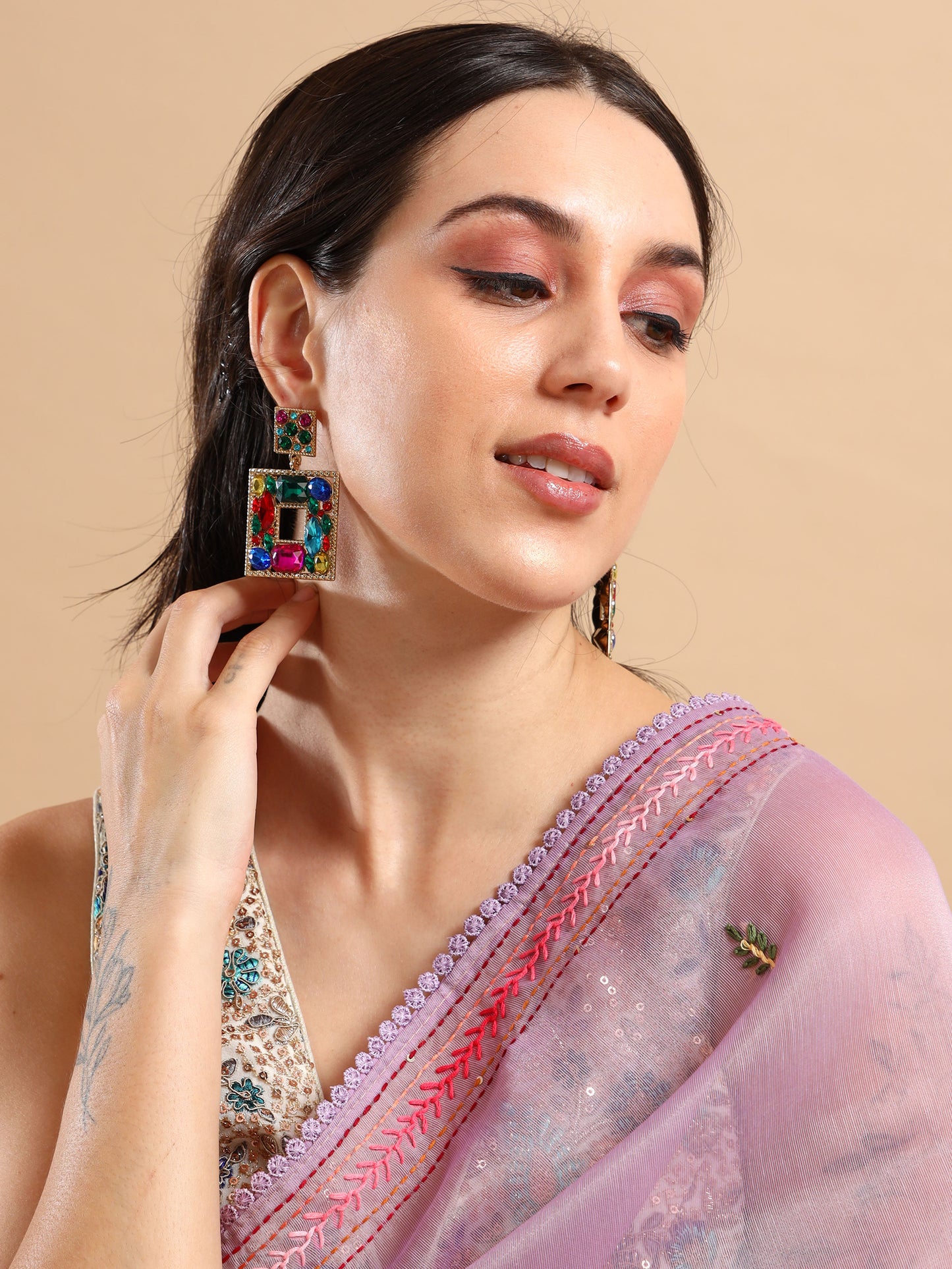 A woman in a light purple saree with pink embroidery looks to the side, touching her neck. She wears bold Esmeralda Earrings and pink eye makeup, with her dark hair tucked behind her ear.
