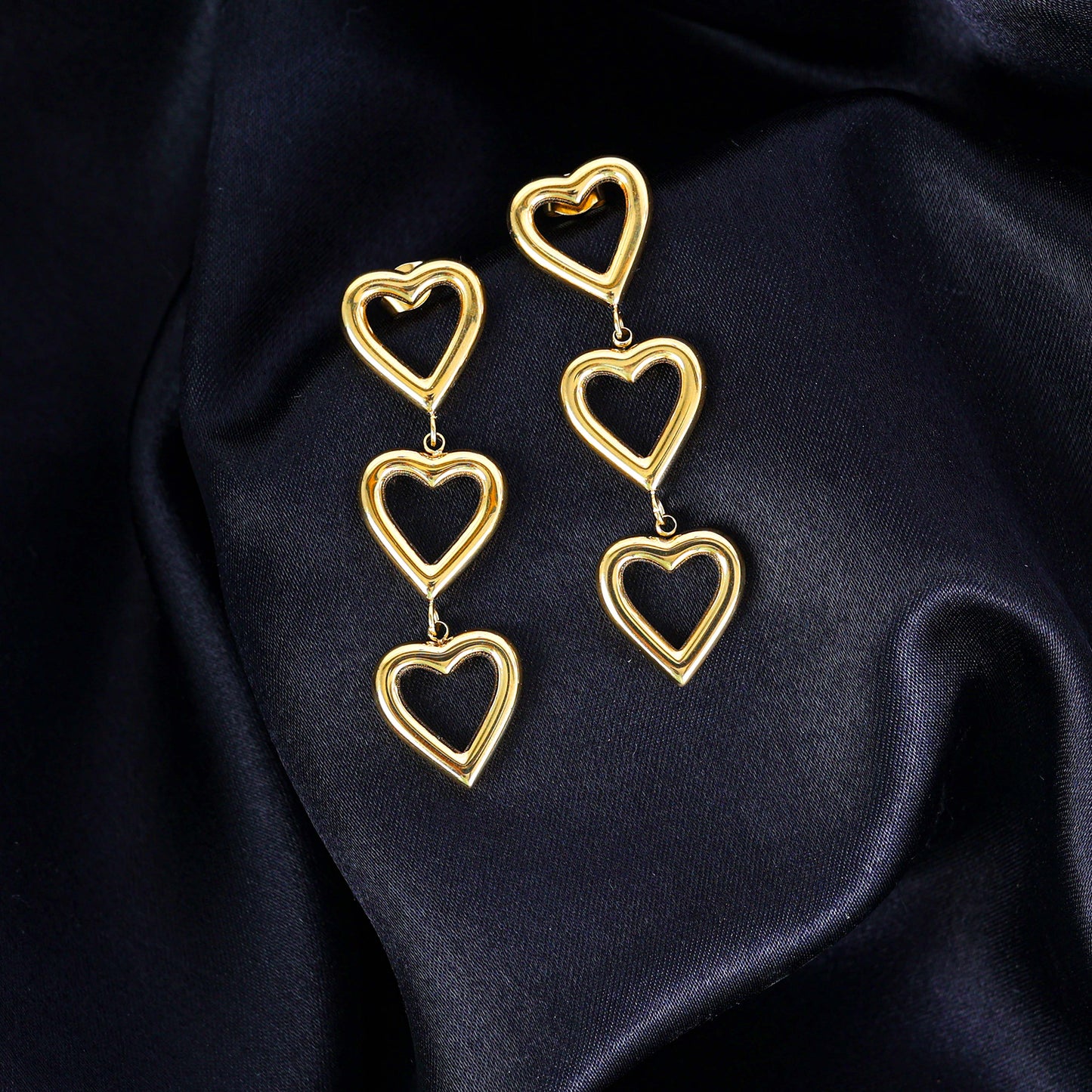 Gold Heart Dangler Earrings featuring three linked open hearts are elegantly showcased on smooth, dark navy blue satin fabric.