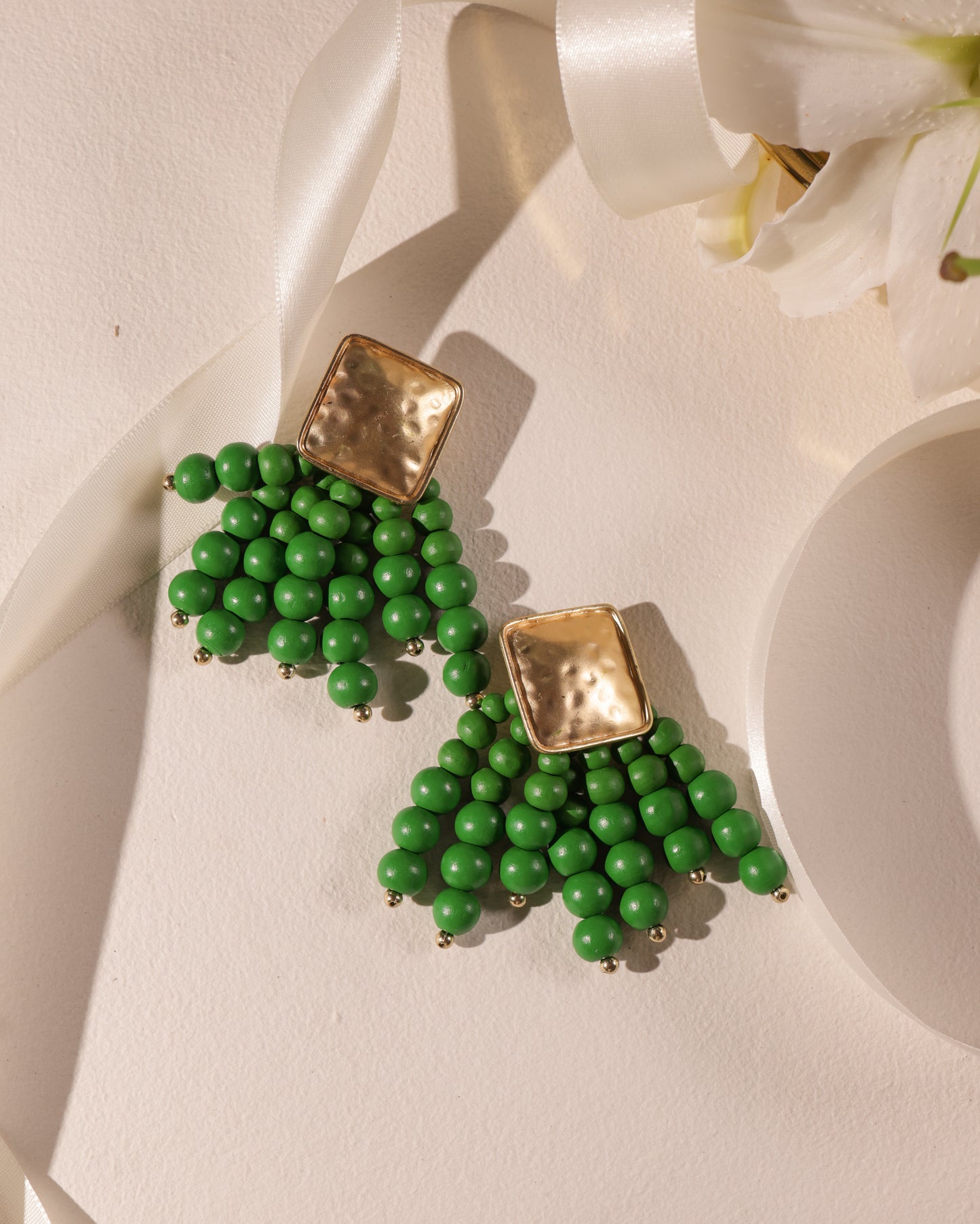 Beady Green and Gold Earrings feature gold-plated, textured square studs with fan-shaped clusters of green beads—displayed on a light surface near a white ribbon and flower—making them perfect vibrant summer jewelry.