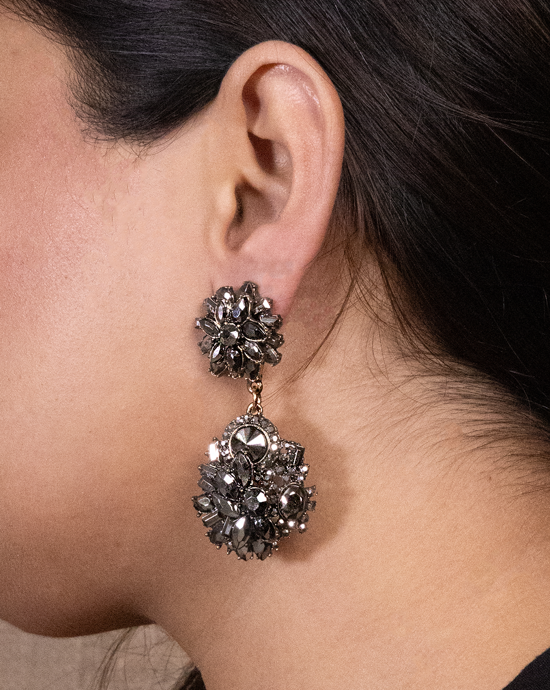 A close-up shows a person wearing Black Magic Earrings—large, ornate double-drop earrings featuring lab-created stones in a floral design. The earrings stand out beautifully against the person’s dark hair and skin.