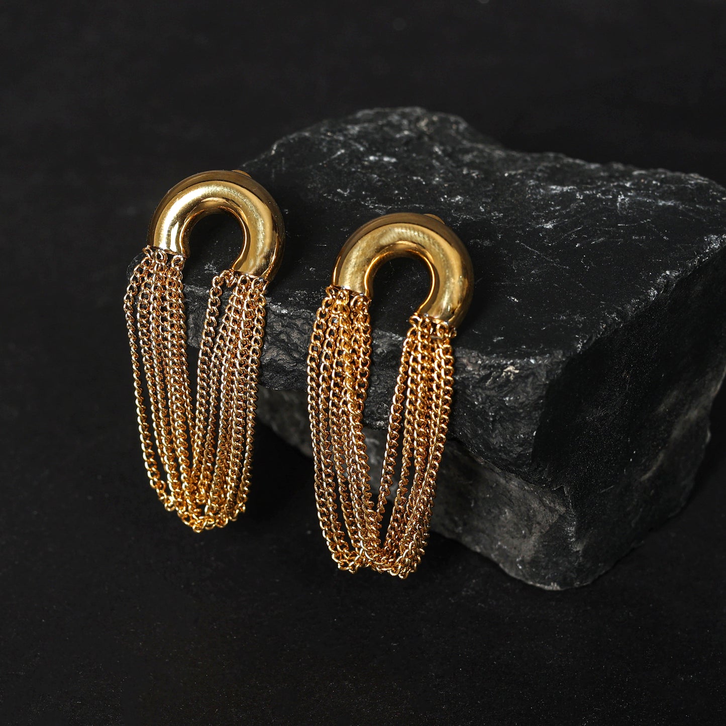 Gold chain earrings featuring thick, gold-tone horseshoe-shaped tops and multiple delicate chain tassels, showcased on textured black stone against a dark backdrop—a timeless fashion accessory.