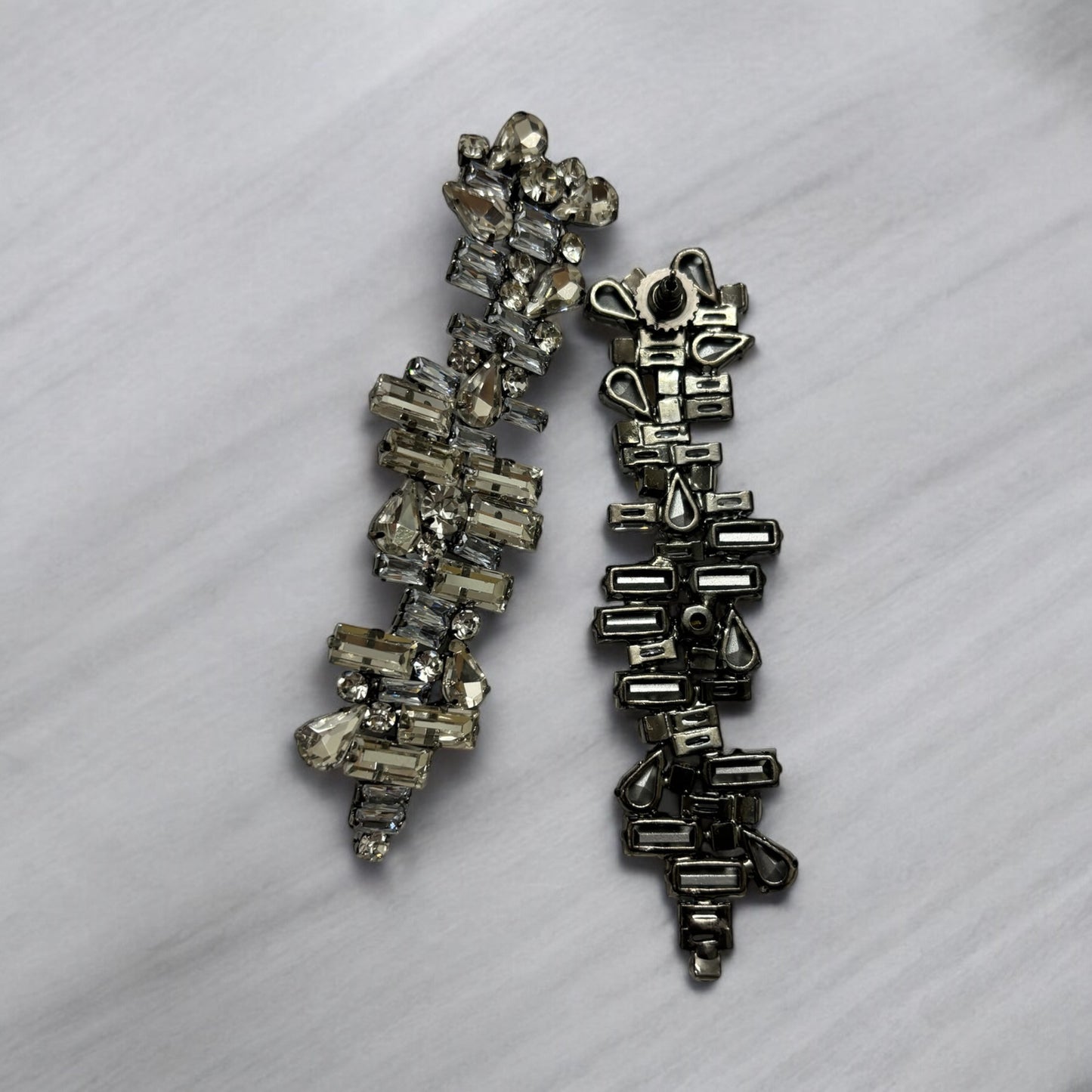 Two metallic crystal earrings rest side by side on a light gray marble surface. The left earring sparkles with clear crystals, while the right displays its mixed metal backing and glossy metallic edge without stones.