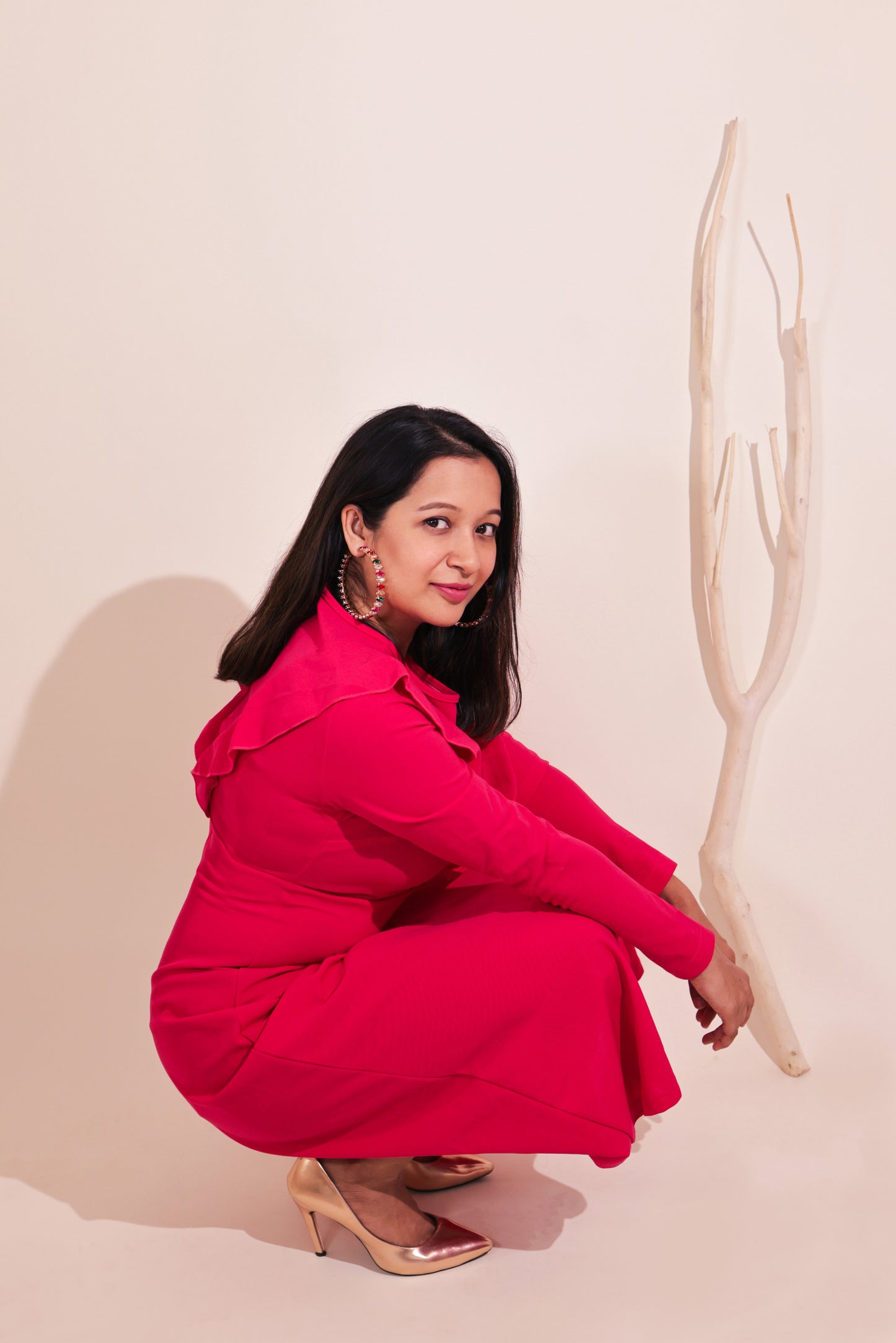A woman in a bright pink dress and metallic heels squats beside a light branch against a neutral background, radiating affordable luxury as she smiles at the camera, showcasing the Big Multicolour Hoops.