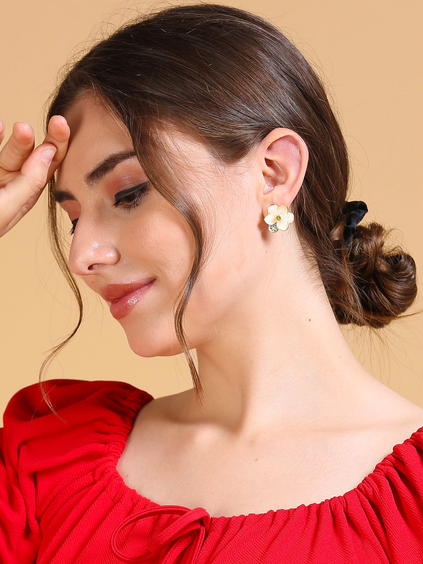 A woman with brown hair in a low bun wears a red top and stylish enamel flower earrings, posing with her hand near her forehead against a beige background.