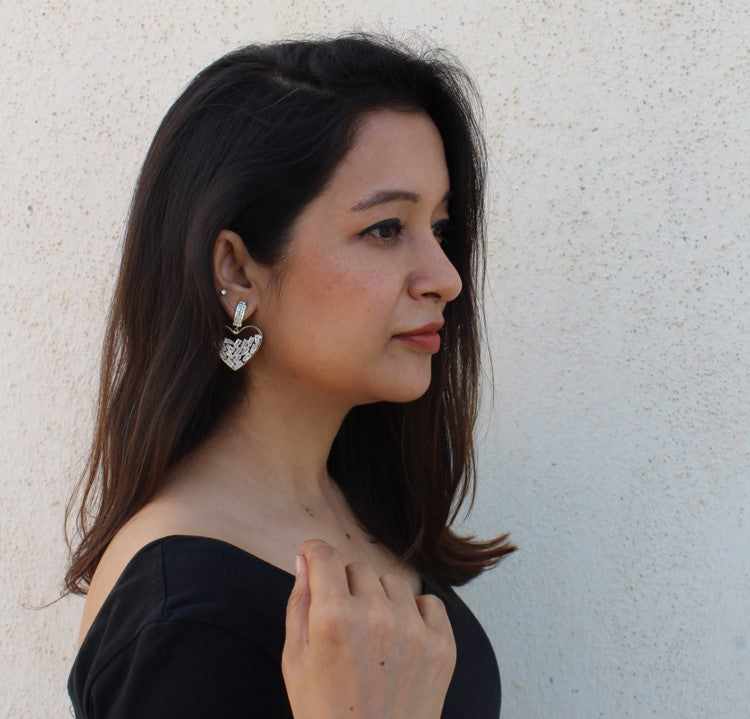 Woman with long dark hair wears a black off-shoulder top and the Hollow Hearts Earrings—large, heart-shaped luxury jewelry with lab-created stones—alongside a small stud. She stands before a light textured wall, looking slightly right.