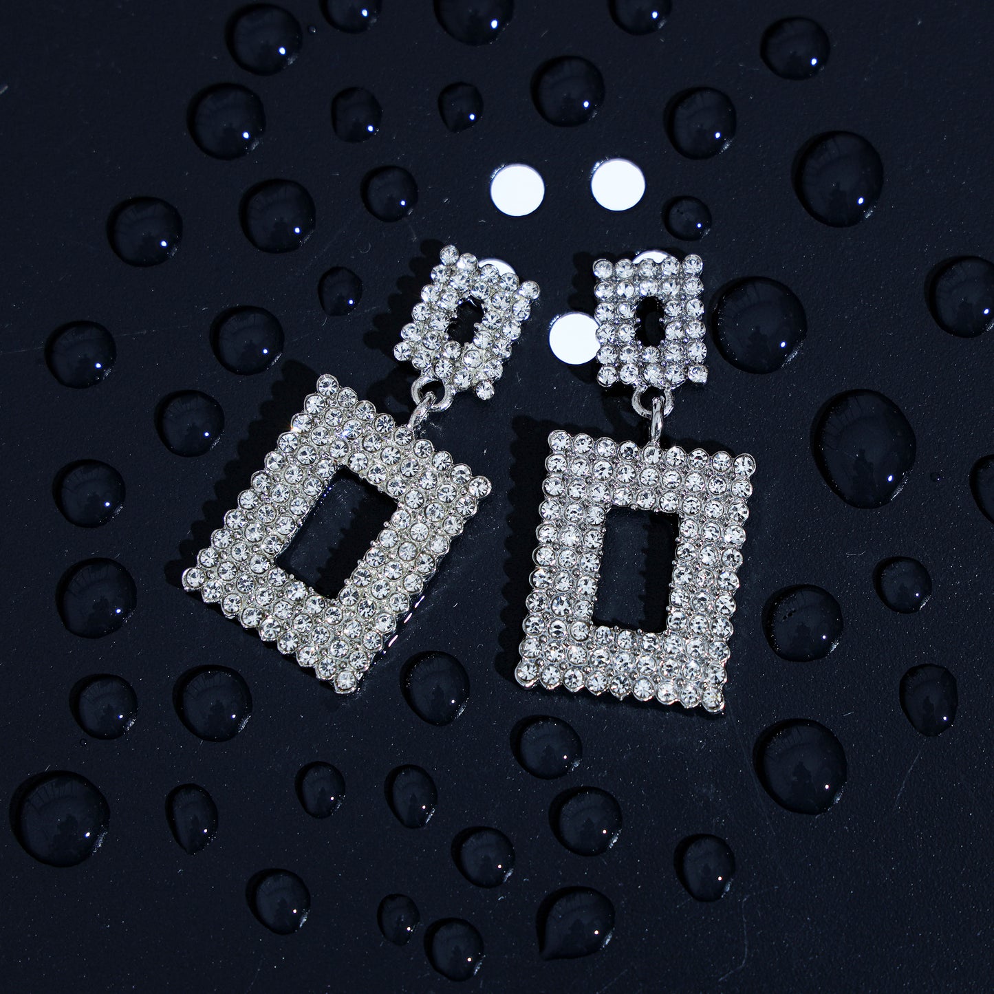 Geometric rhinestone earrings in silver with a rectangular design are displayed on a dark surface with scattered water droplets, highlighting their luxurious appeal.