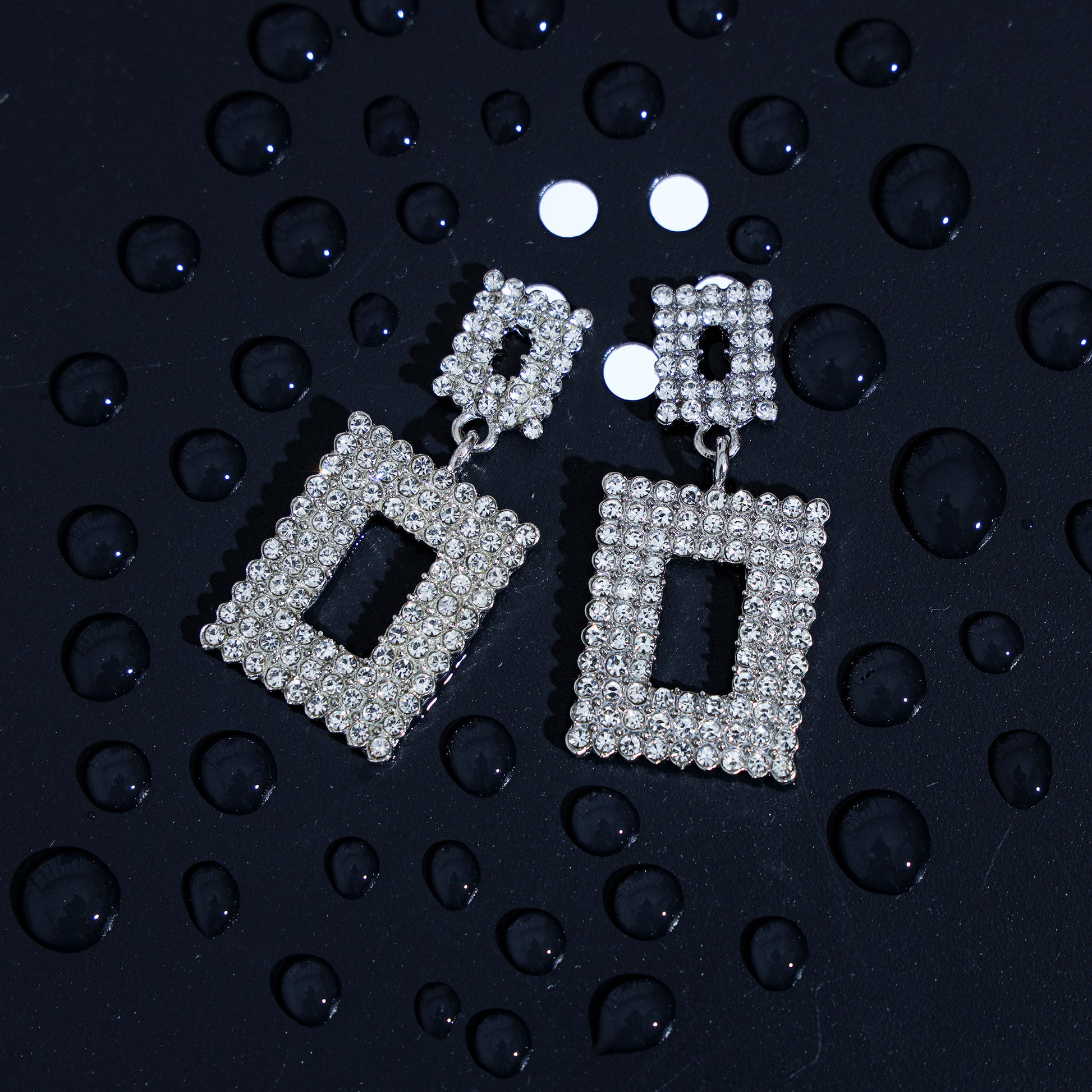 Geometric rhinestone earrings in silver with a rectangular design are displayed on a dark surface with scattered water droplets, highlighting their luxurious appeal.