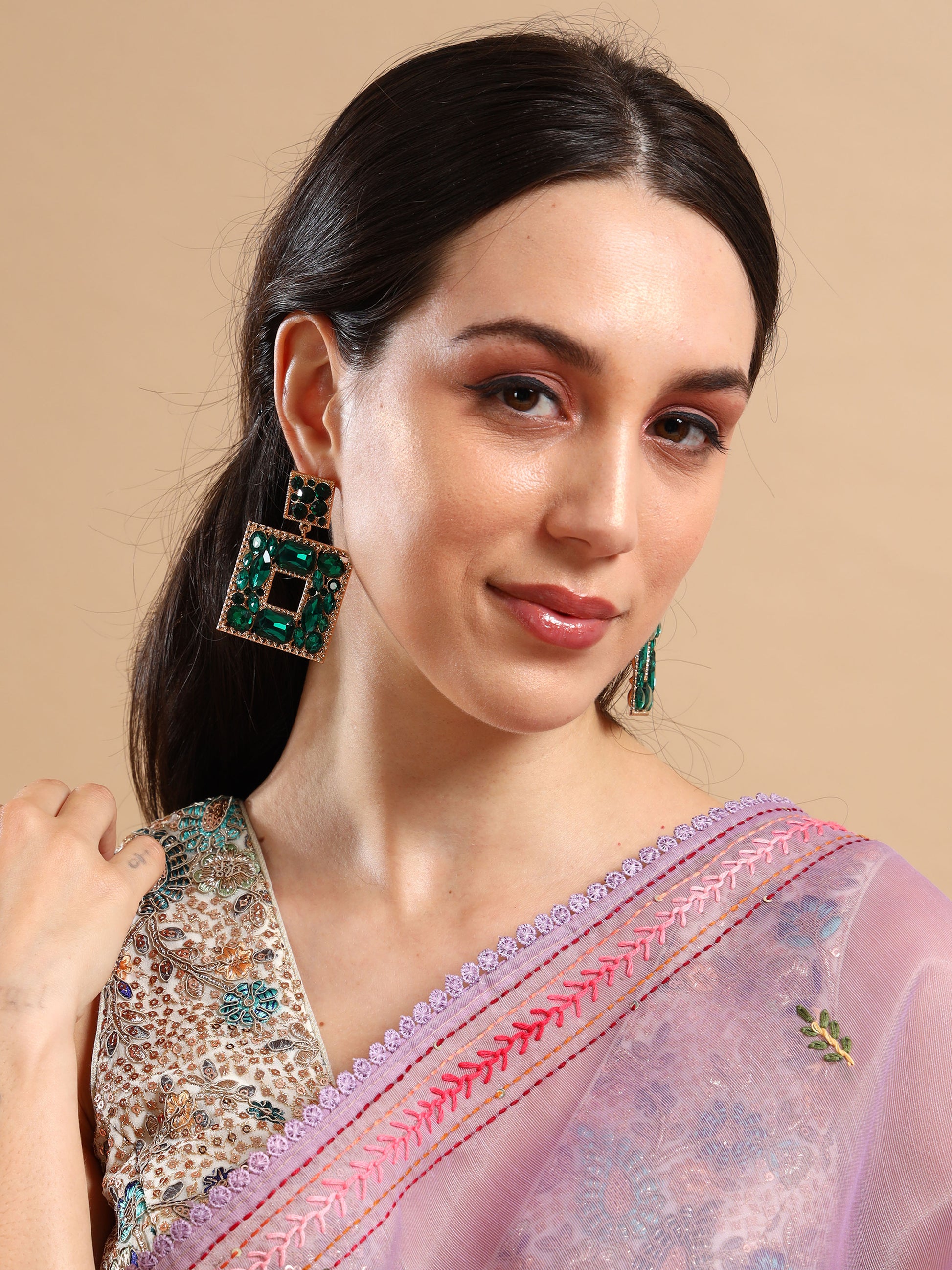 A woman with dark hair in a low ponytail wears large Green Goblin Earrings and a lavender saree with intricate embroidery, smiling softly against a beige background.