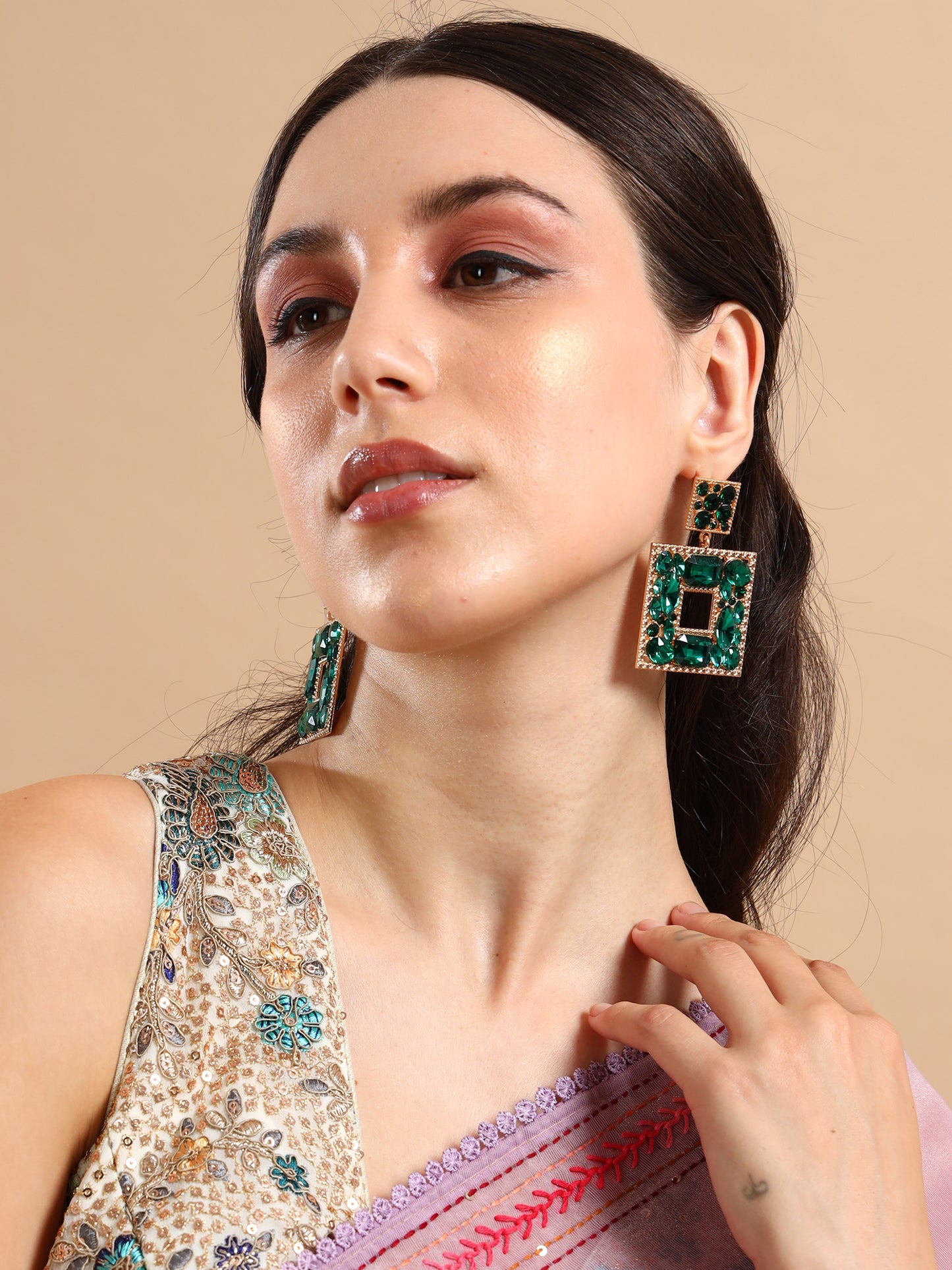 A woman in a sequined blouse and lavender saree models the Green Goblin Earrings—bold, green and gold-plated square statement pieces. With long dark hair and subtle makeup, she gazes upward, her hand softly resting near her neck.