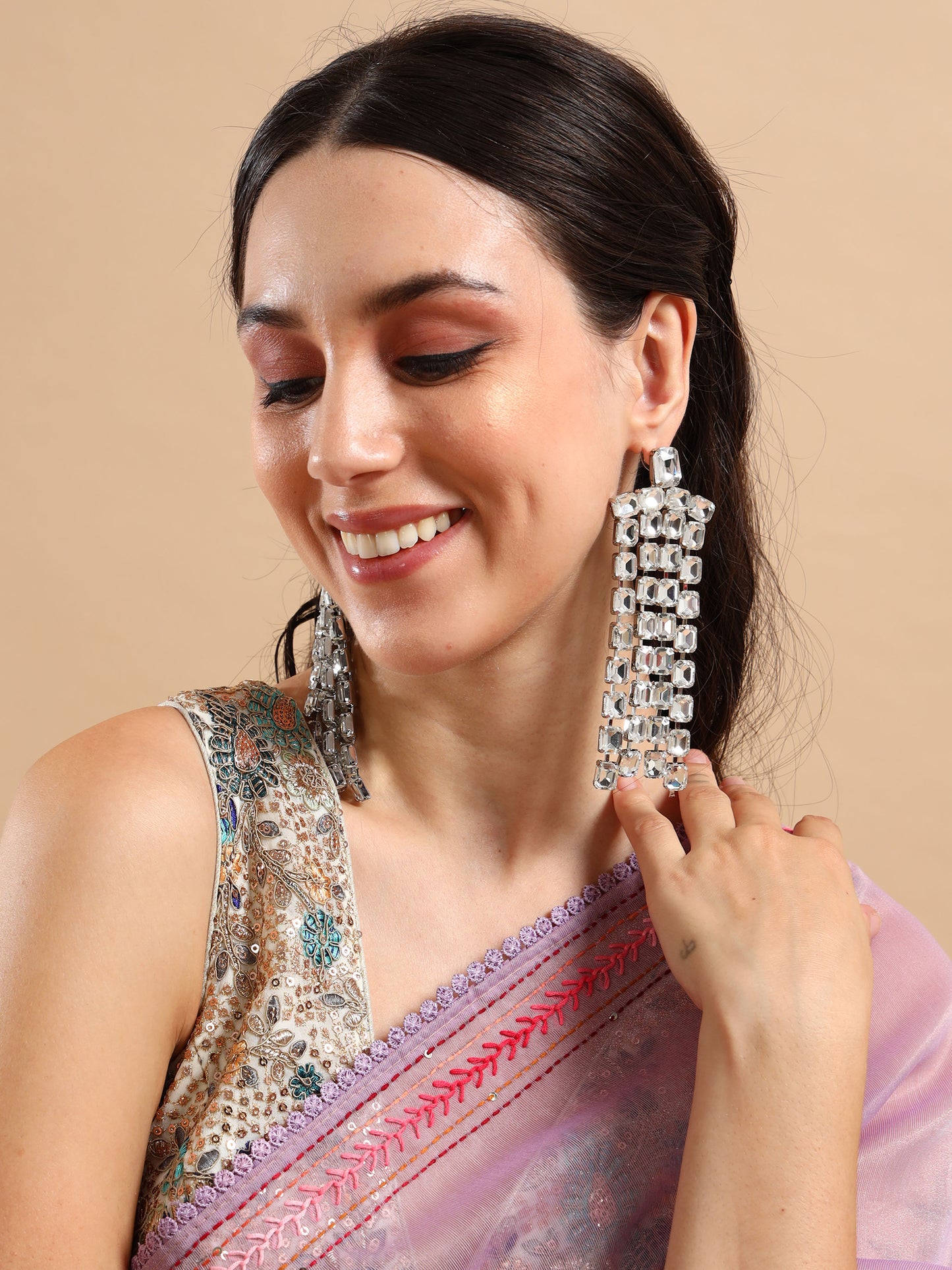 A woman wearing a colorful, embroidered saree with a lavender border smiles while showcasing Long Rhinestone Tassel Earrings. Her bold eyeshadow complements her look as she poses against a beige background.