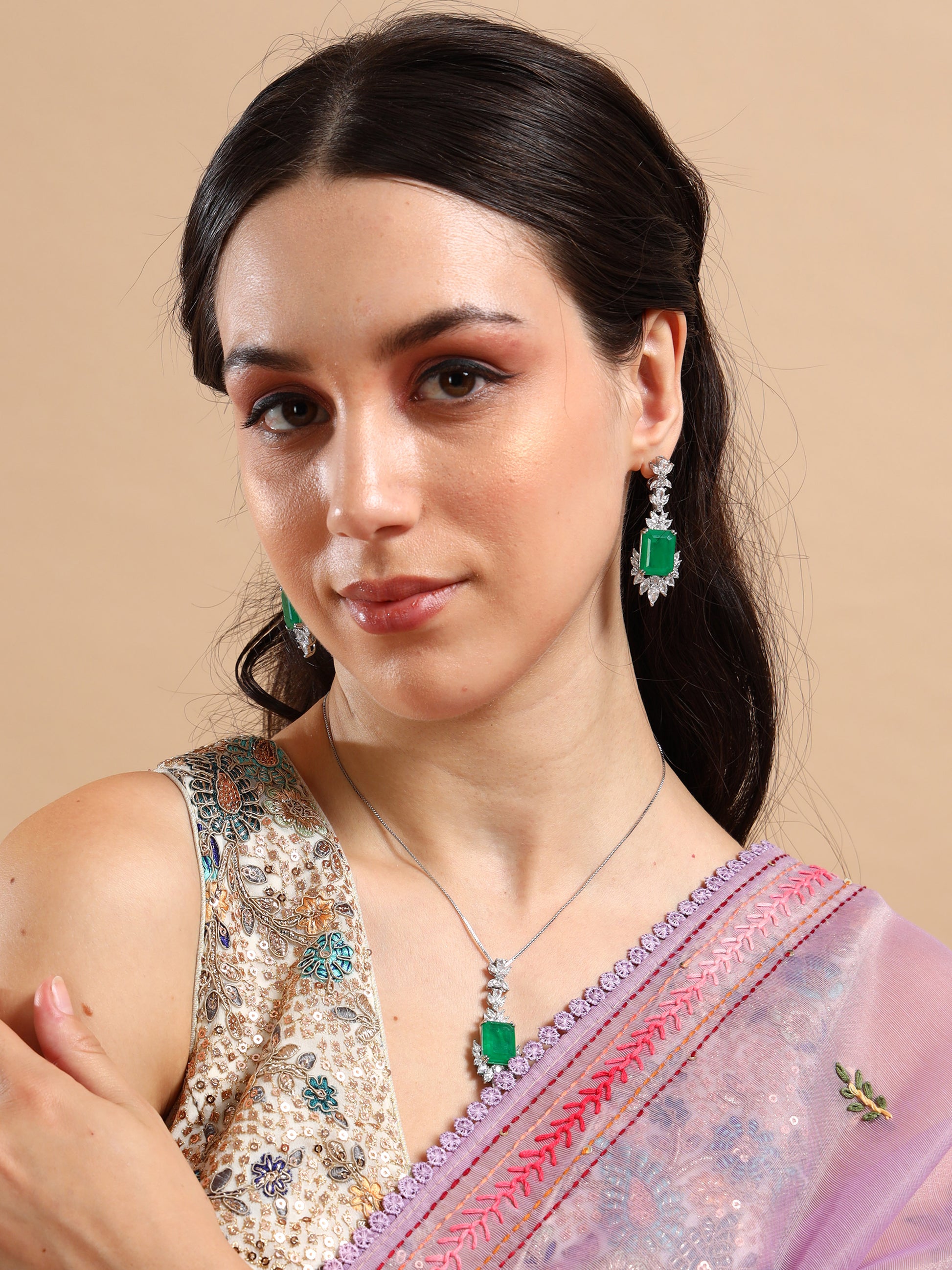 A woman in a sequined blouse and lavender saree with a pink border, accessorized with the Queen’s Earrings Set and a green stone necklace, poses against a beige background.