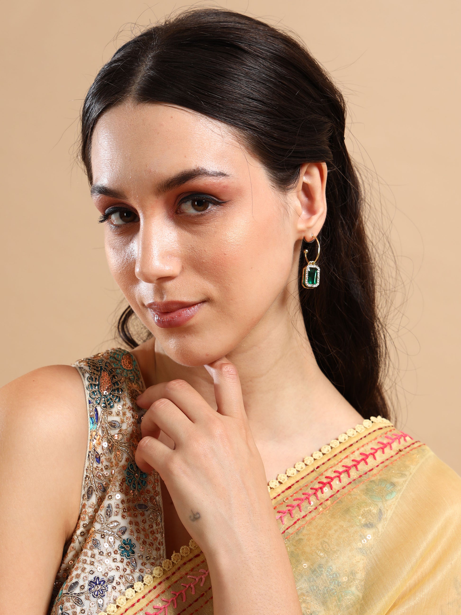 A woman with long dark hair, wearing a yellow sari with pink trim and a sequined blouse, poses against a beige background. She touches her chin while showcasing luxury jewelry: elegant green two layer earrings.