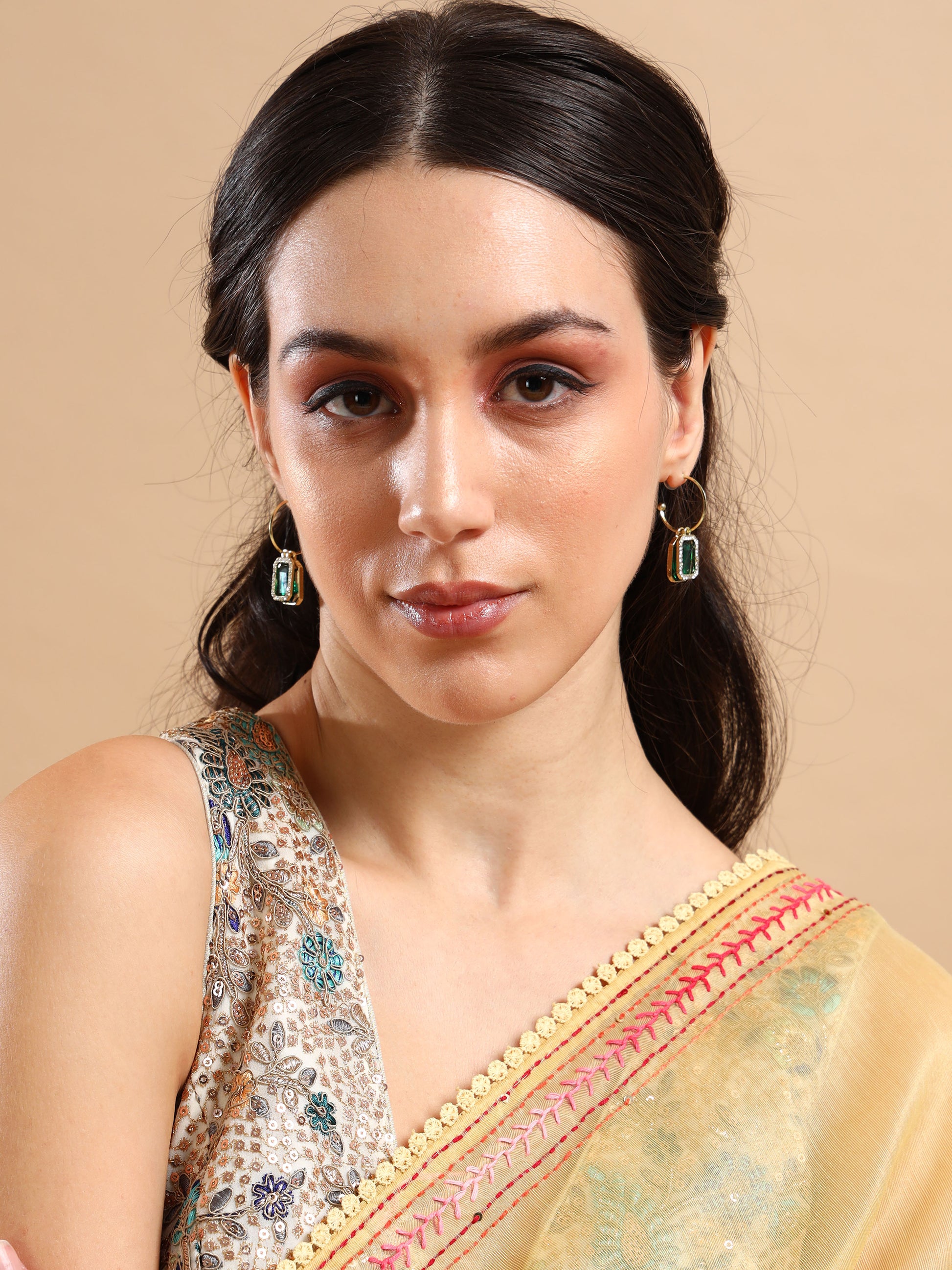 A woman with long dark hair wears Green two layer earrings featuring green lab-created stones and a beige saree with a red and gold border, posing against a tan background. Her makeup is neutral, and she has a calm expression.