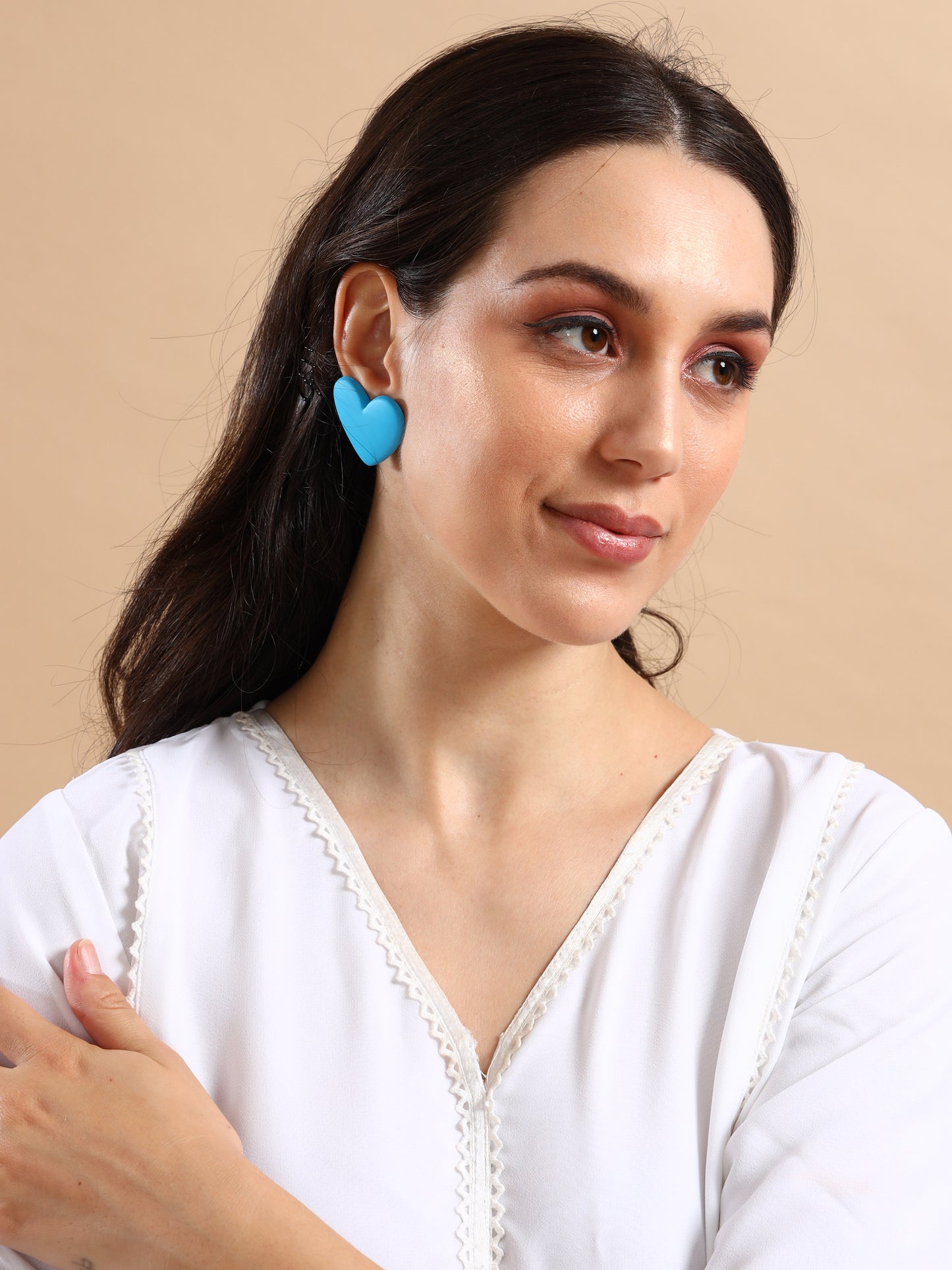 A woman with long dark hair, wearing a white blouse and the Love Hearts Earrings—large blue heart-shaped gold plated earrings—poses against a beige background, looking slightly to the side with a gentle smile.