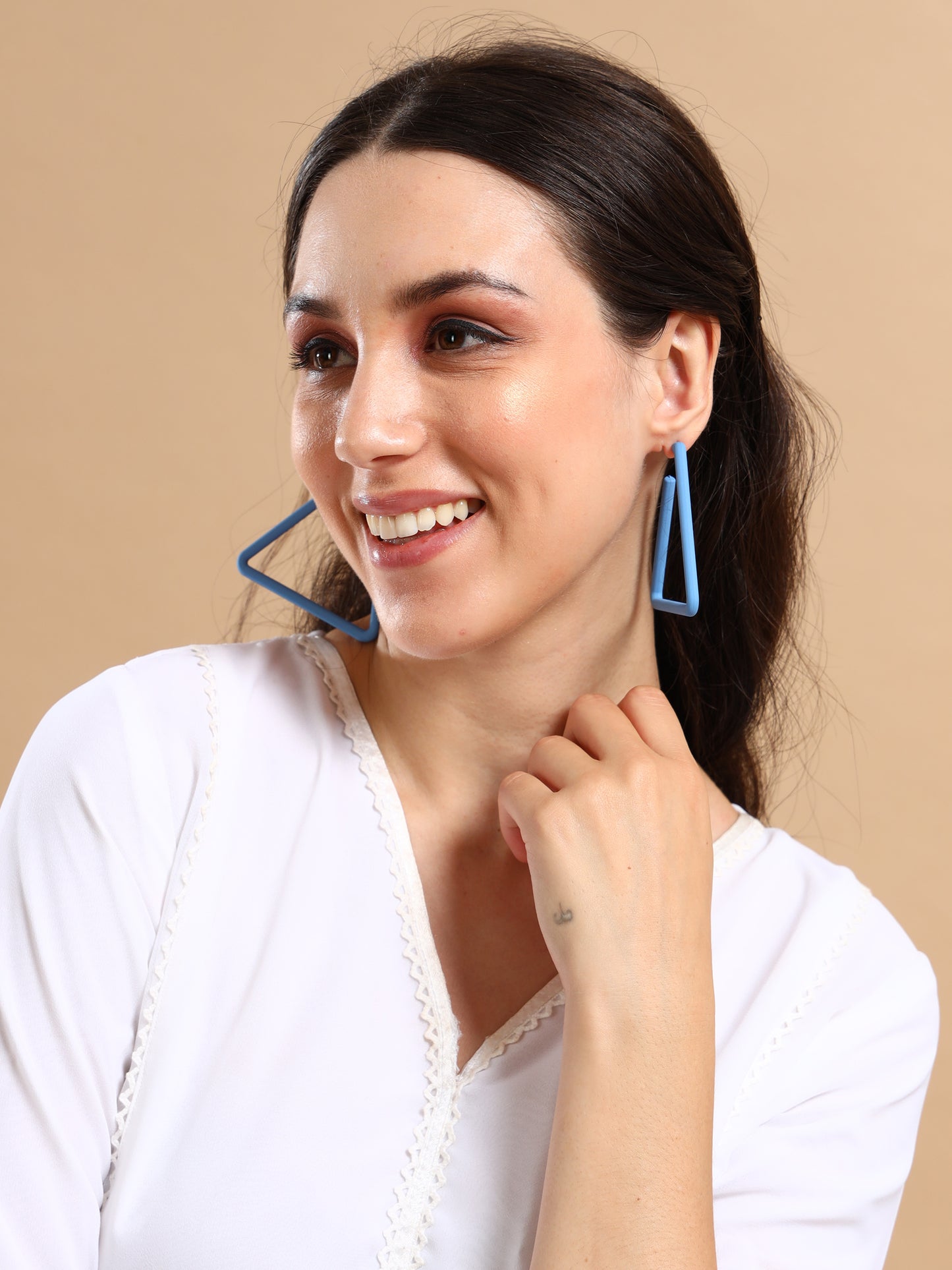 A smiling woman with long dark hair wears our Triangle Earrings—18K gold plated and affordable—posing in a white top against a beige background, radiating effortless luxury as she touches her chin.