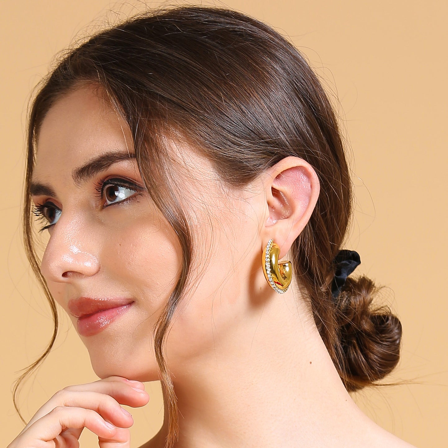 A woman with light skin and brown hair in a low bun wears a red off-shoulder top and Half Circle Hoop Earrings. She gazes to the side, gently touching her chin, set against a beige background.