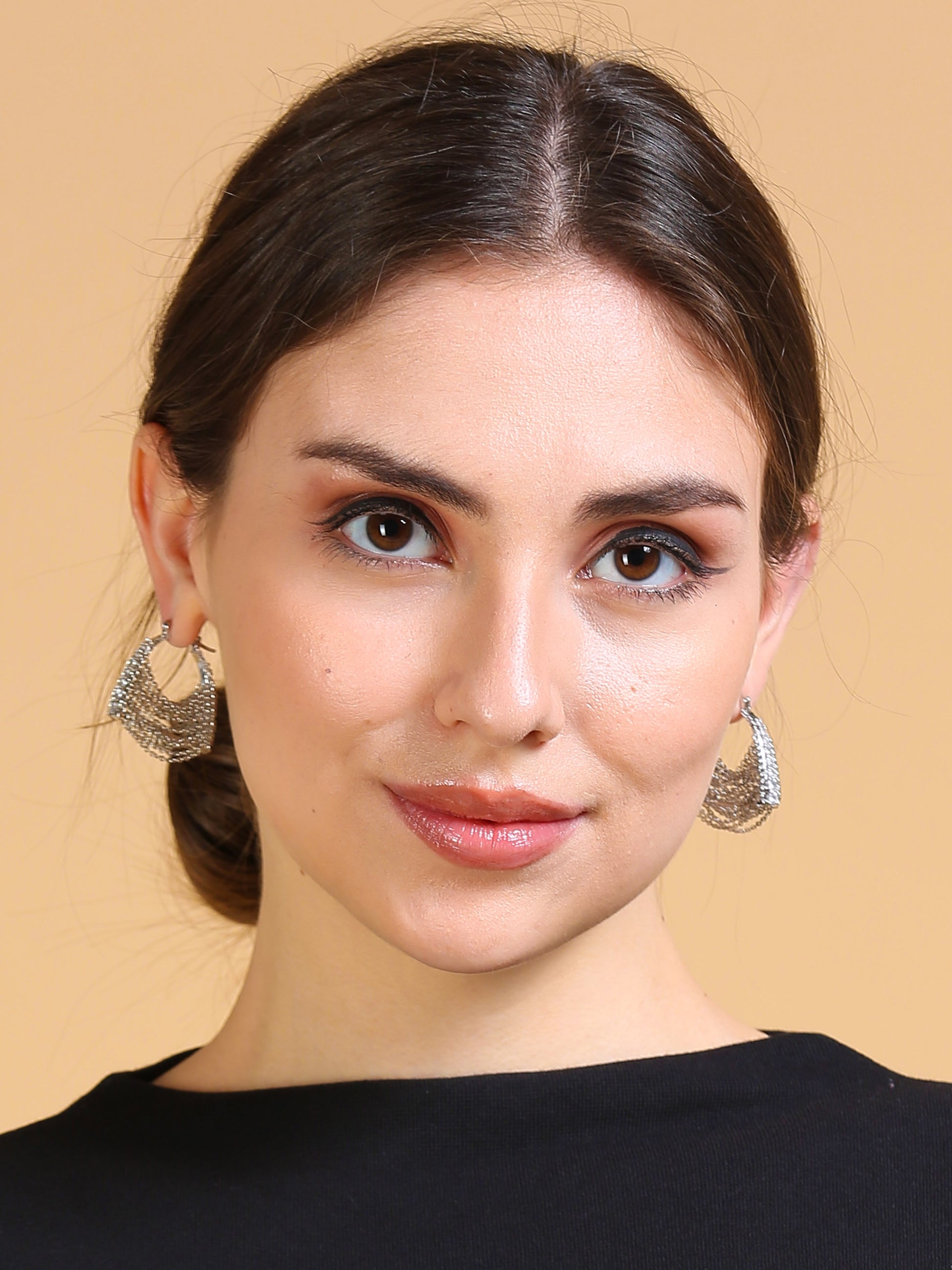A young woman with brown hair in a low bun, wearing Silver Chain Bag Earrings and a black top, poses against a peach background with a soft smile—her lightweight jewelry pouch keeps her striking earrings safe.
