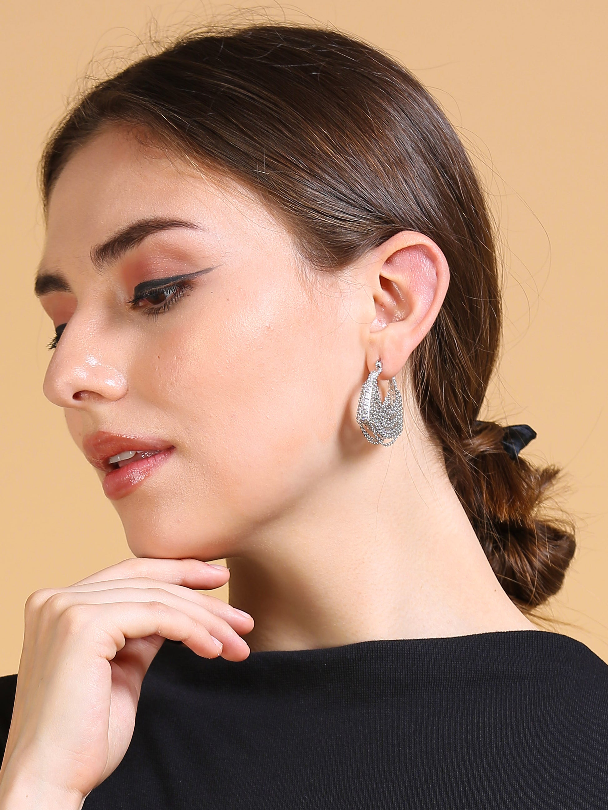 A woman with brown hair in a low bun, wearing a black top and Silver Chain Bag Earrings, looks down thoughtfully with her hand near her chin against a beige background, next to a lightweight jewelry pouch.