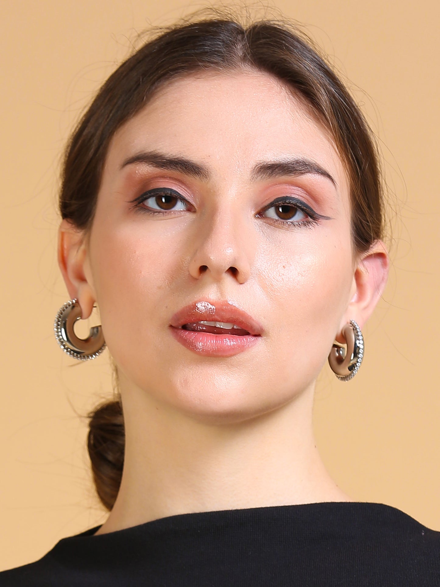 A woman with light skin and brown hair in a low bun, wearing glossy nude lipstick, subtle eye makeup, Half Circle Silver Hoop earrings, and a black top poses against a plain beige background.