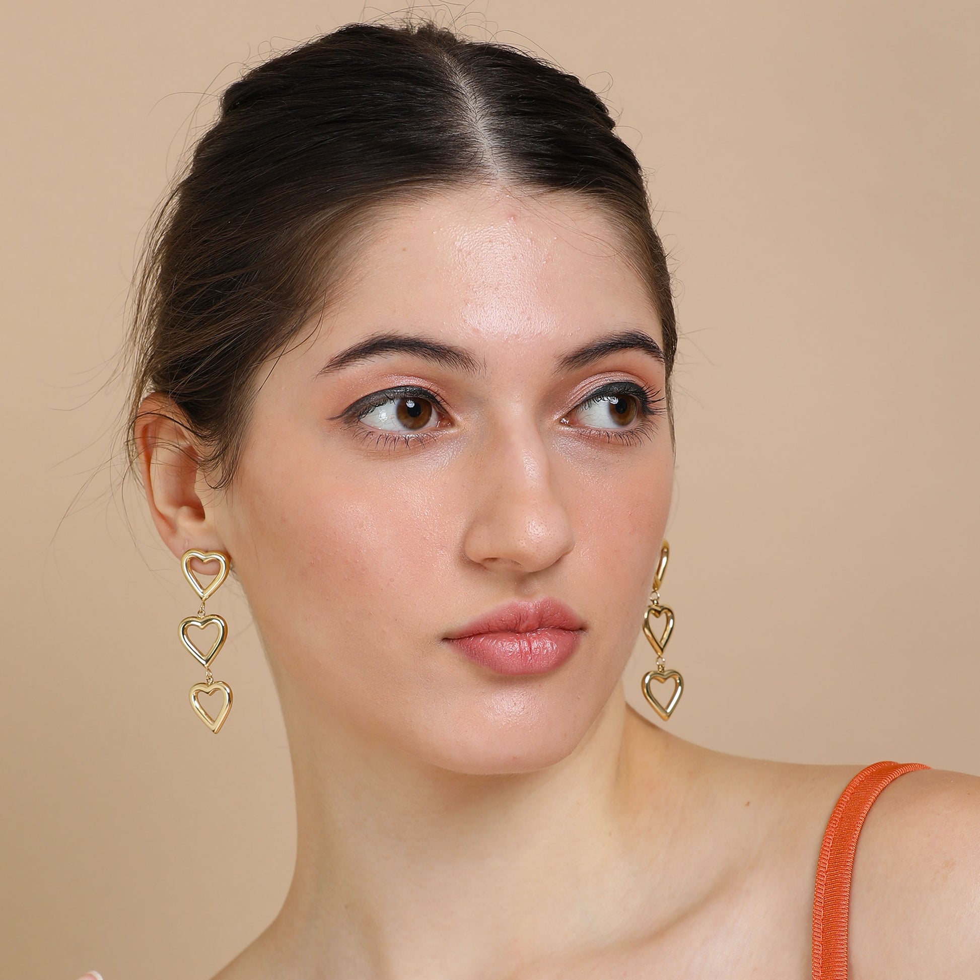 A woman with fair skin and dark hair tied back wears Gold Heart Dangler Earrings and an orange strap top, gazing slightly to the side against a beige background.
