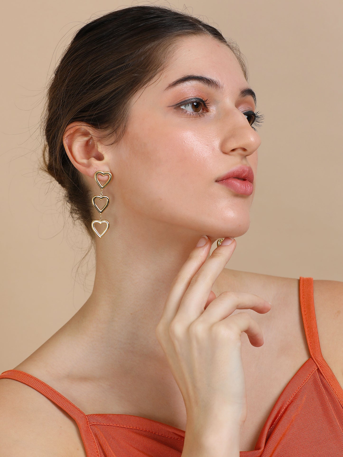 A woman in gold heart dangler earrings poses with her hand by her chin, gazing to the side against a beige background.