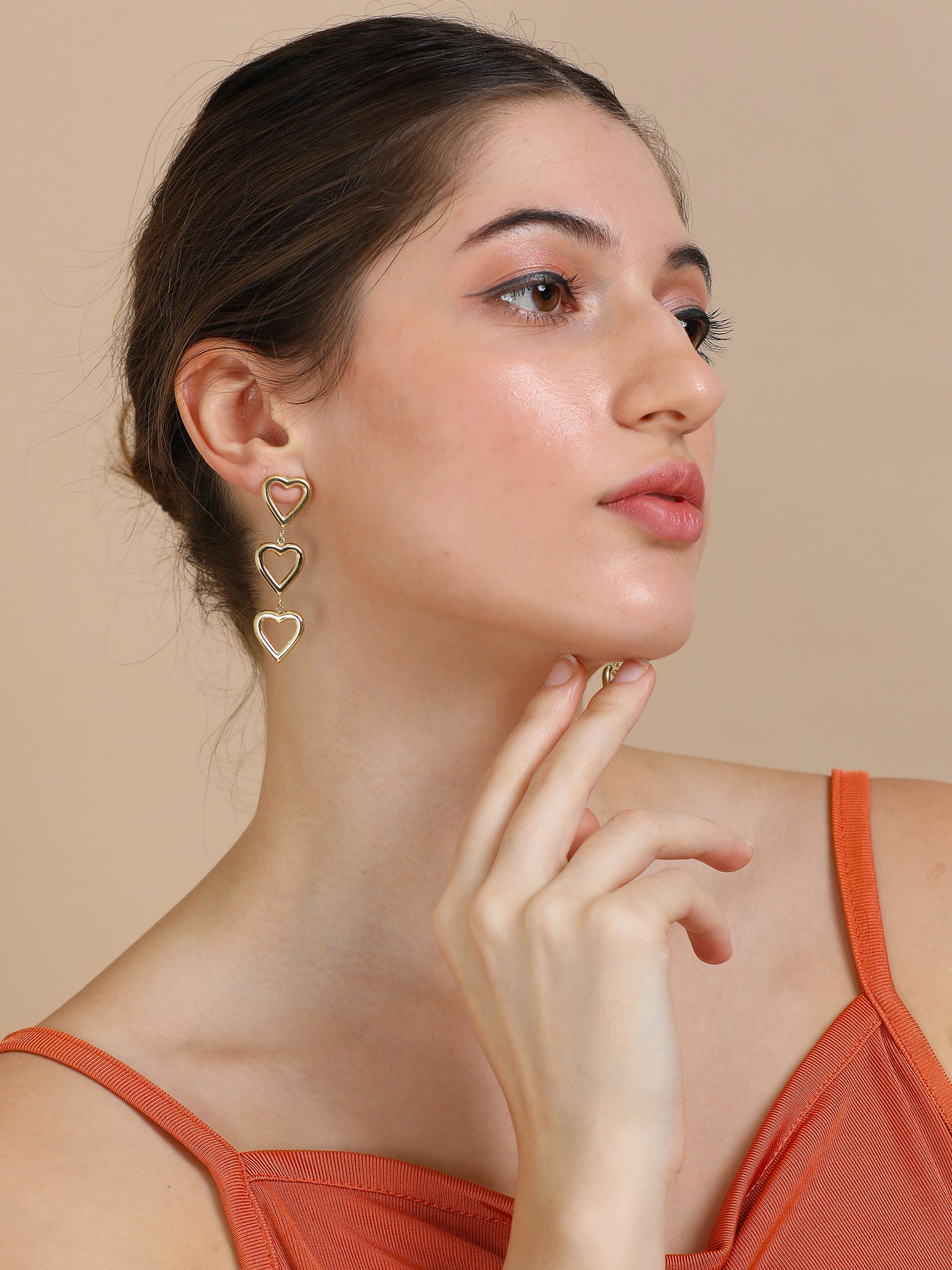 A woman in gold heart dangler earrings poses with her hand by her chin, gazing to the side against a beige background.