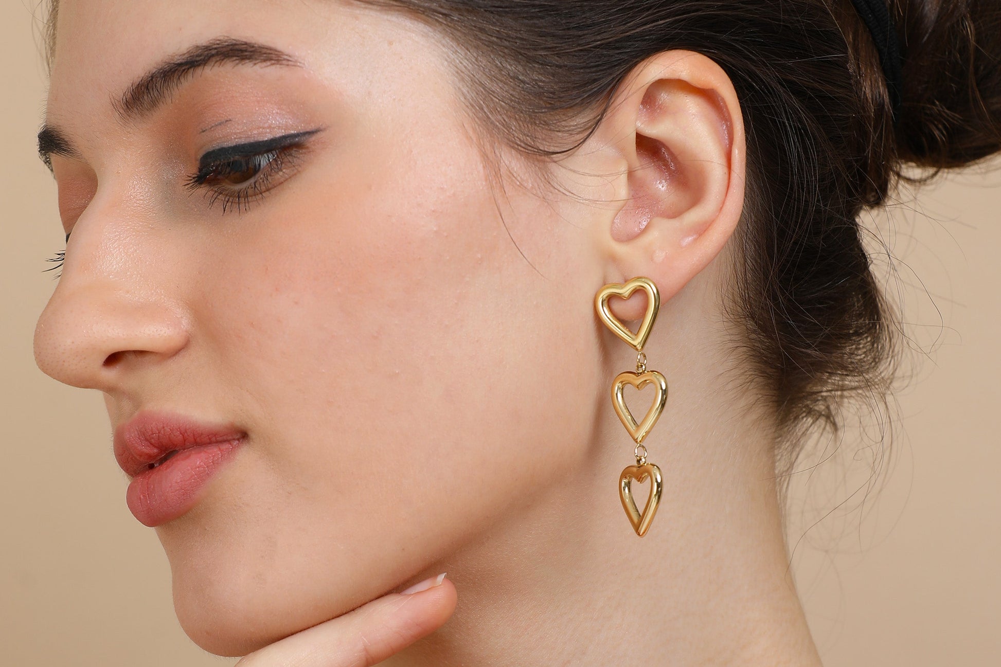 A woman with brown hair in a bun wears Gold Heart Dangler Earrings. She is dressed in an orange top, looks to the side, touches her chin, and poses against a beige background.