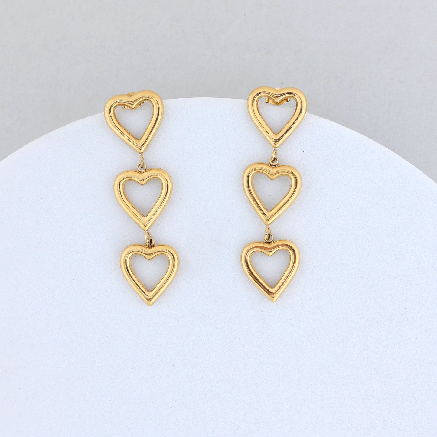 The Gold Heart Dangler Earrings feature three linked open heart shapes in each earring, beautifully displayed against a light grey and white background.