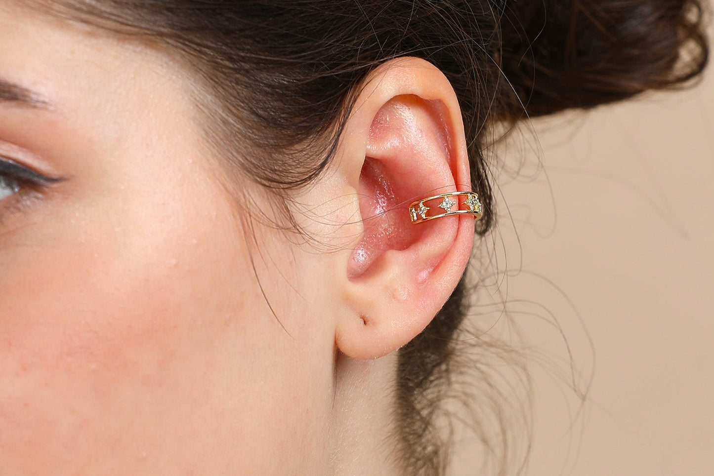A close-up shows a woman's ear adorned with the Small Gold Ear Cuff featuring star-shaped cutouts. Her hair is tied back against a soft beige background, making this elegant, gold plated piece ideal for stylish ear cuff enthusiasts.