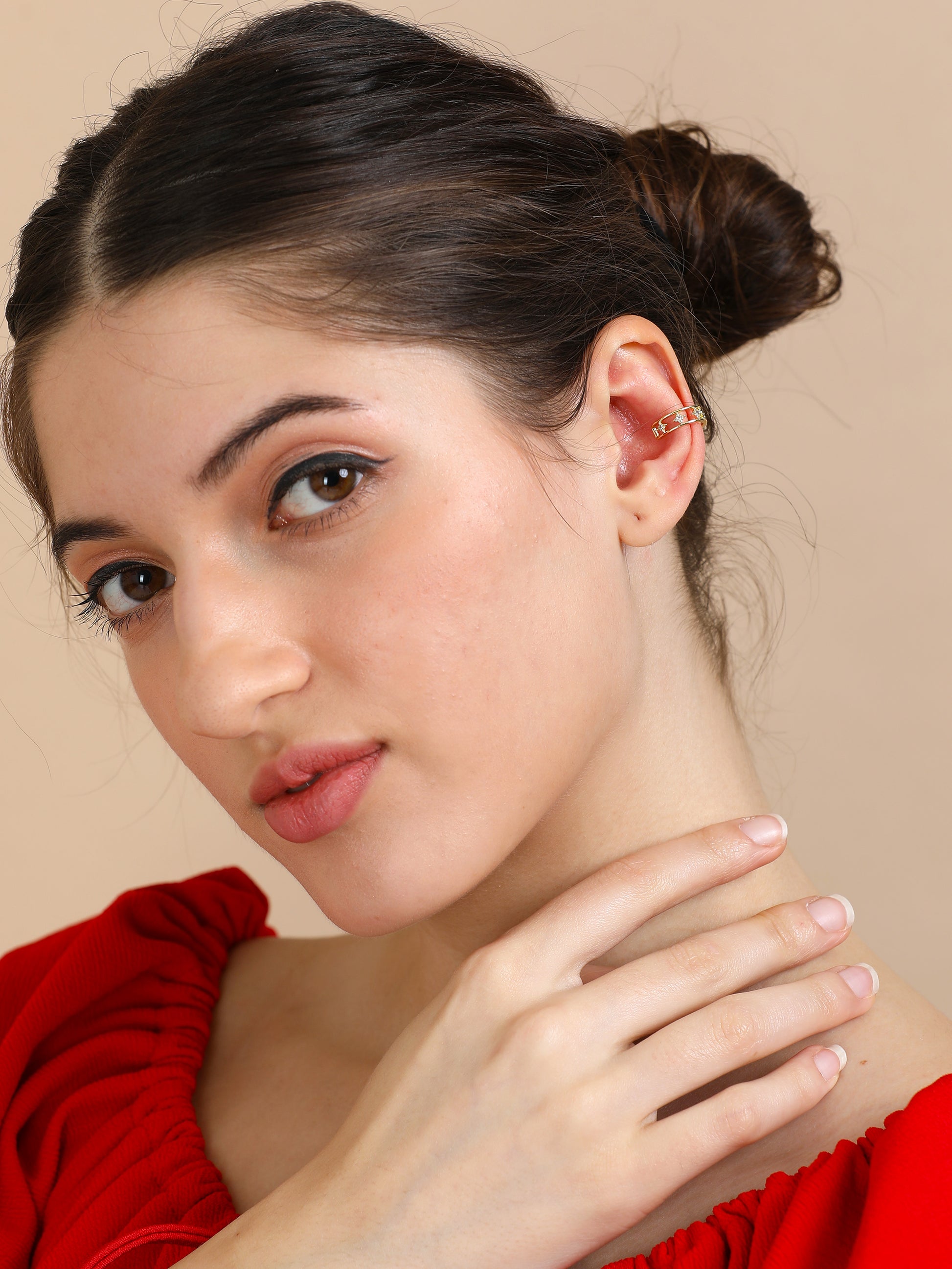 A woman with fair skin and dark hair tied back wears a red top and the Small Gold Ear Cuff as she gently rests her hand on her neck, gazing at the camera with a subtle smile against a beige background.