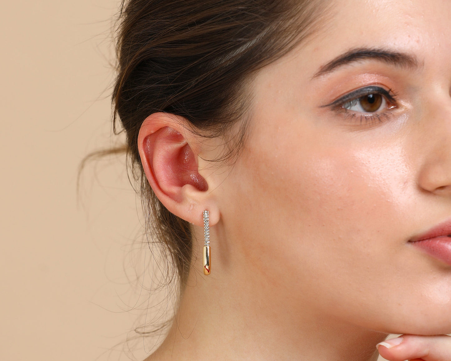 A woman with fair skin and brown hair in a low bun wears a red off-shoulder top and Long Rectangle Gold Hoops, resting her chin on her hand and gazing to the side against a beige background.