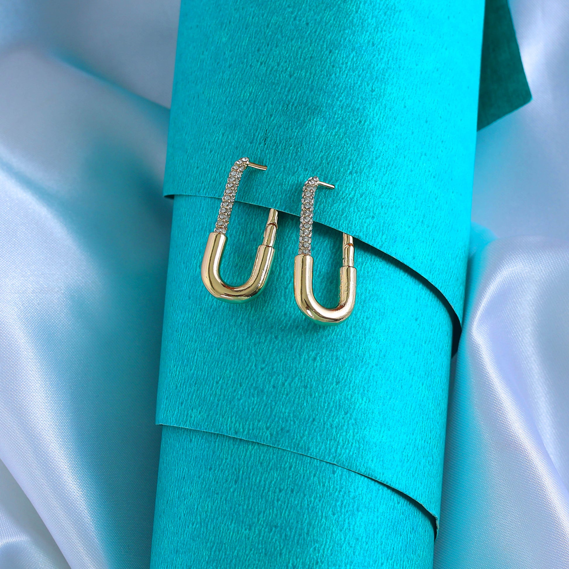 Long Rectangle Gold Hoops with textured upper sections rest on a rolled teal sheet, surrounded by shiny, light blue satin fabric.