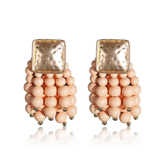 The Beady Peach and Gold Earrings feature textured gold square studs with clusters of round, pale peach beads, each bead accented by a small gold tip. This statement summer jewelry is beautifully reflected on a white surface.