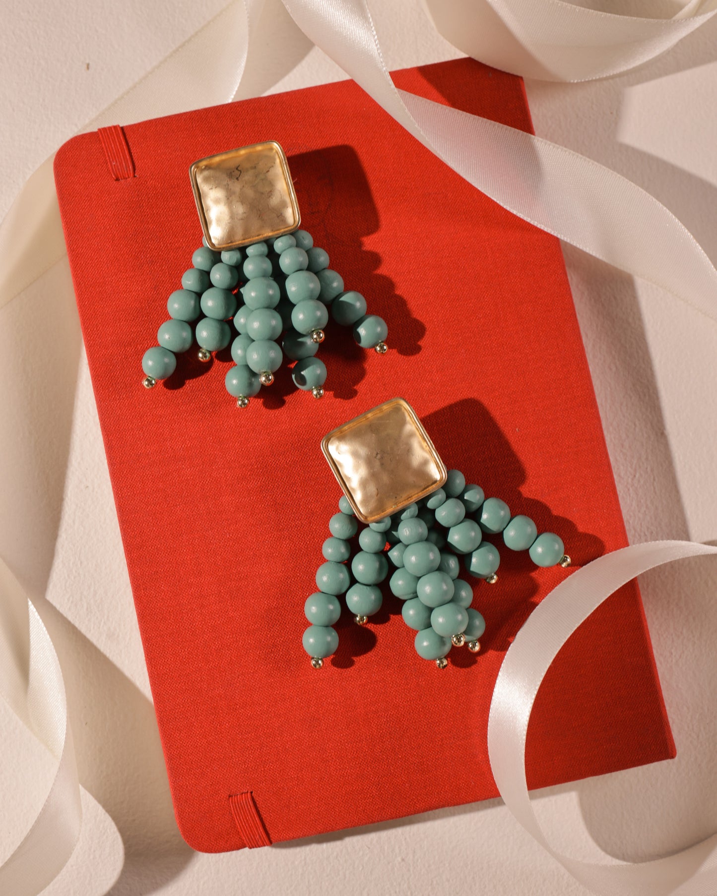 Beady Blue and Gold Earrings are showcased on a closed red hardcover notebook, accented by a loosely draped white satin ribbon.