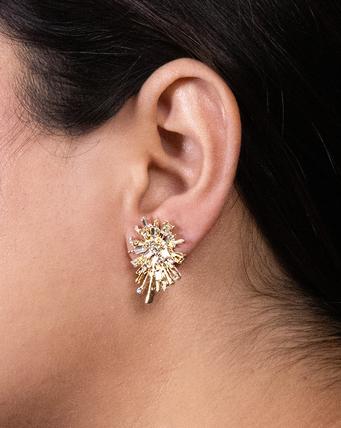 A woman's dark hair is pulled back as she wears Gold Firework earrings—24K gold plated statement pieces with a sunburst design, sparkling lab-created stones, and multiple gold rays.
