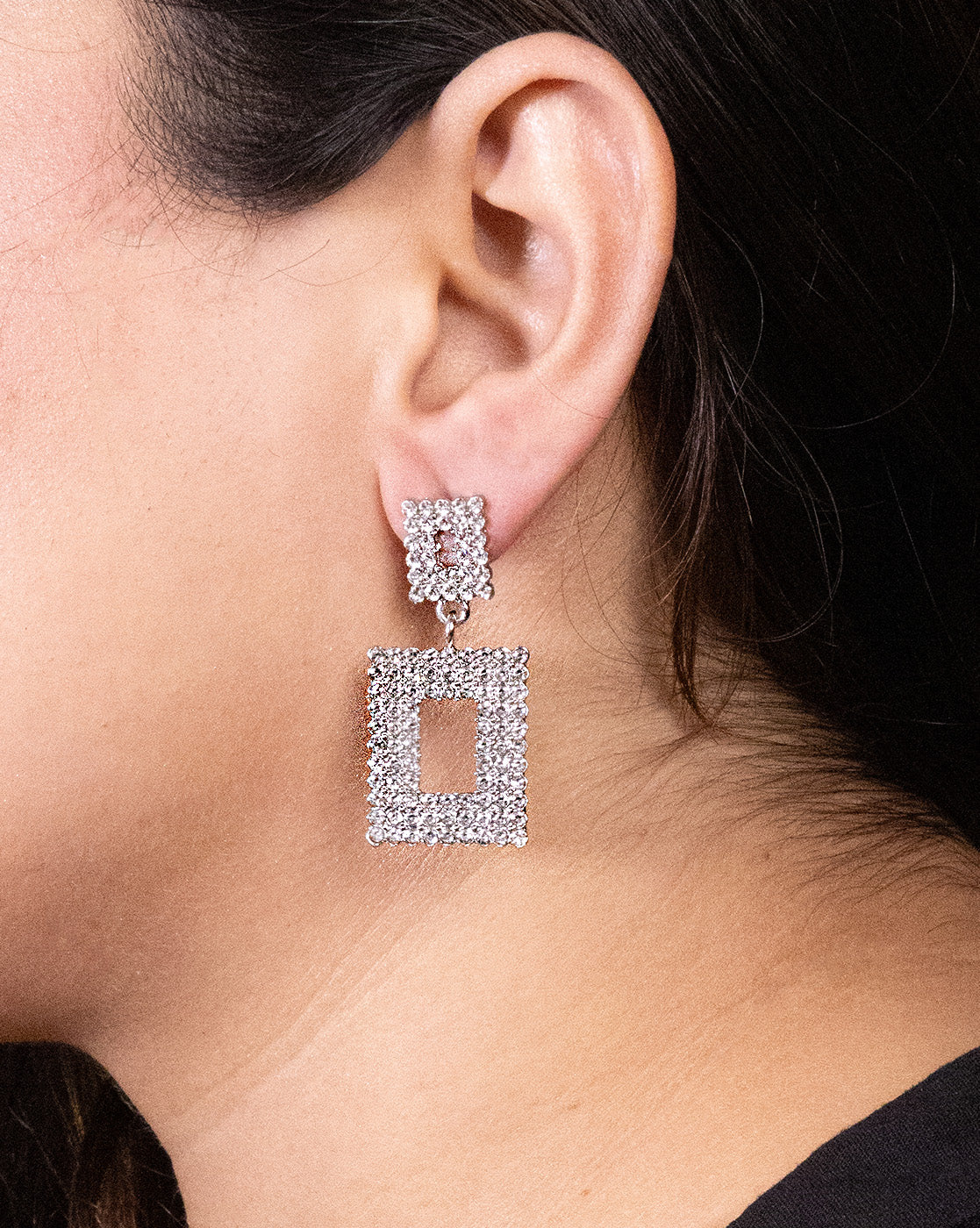 A close-up of a woman's ear shows her wearing Geometric rhinestone earrings—luxurious rectangular drop earrings with a smaller square stud, both covered in clear rhinestones. The background is blurred.