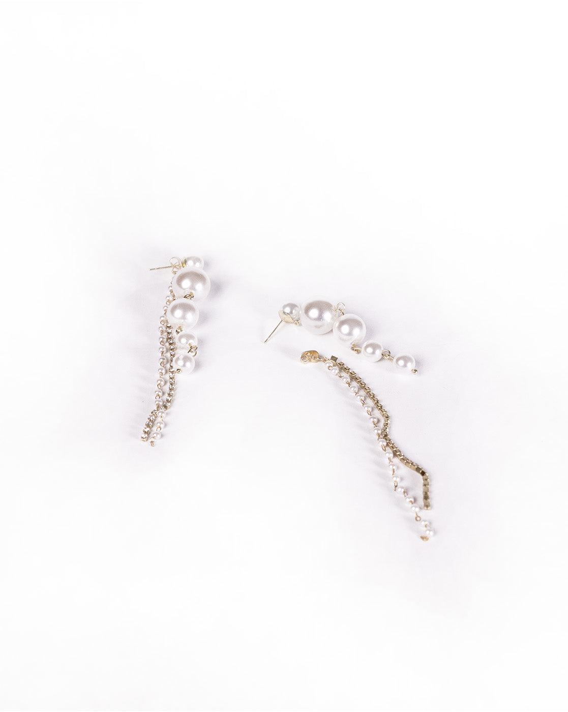 The Pearl Tassel Earrings feature an asymmetrical design with white pearls and gold chains: one earring has three pearls with long tassels, while the other has four pearls with shorter chain accents, all displayed on a white background.