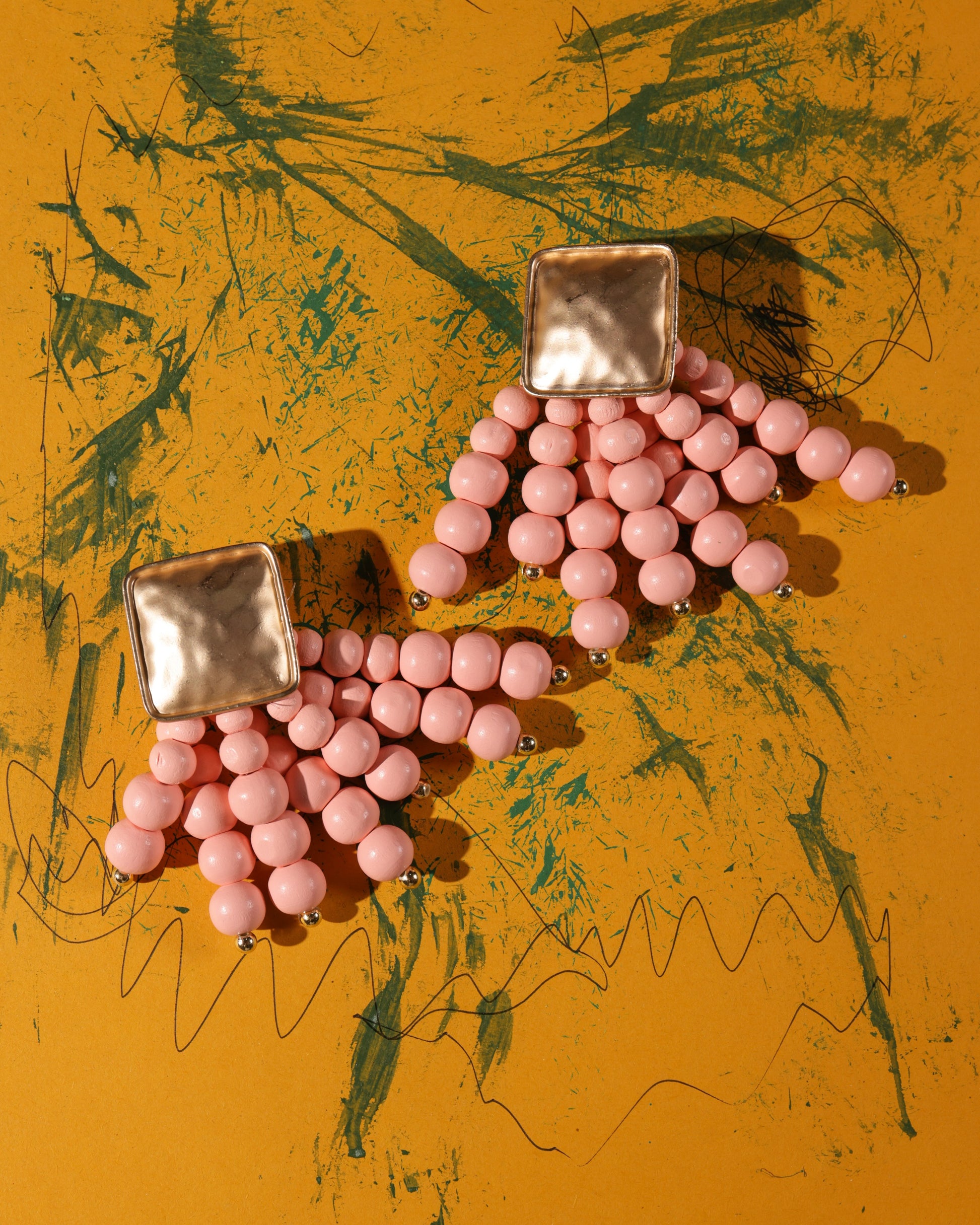 Beady Pink and Gold Earrings: Gold plated earrings with square studs and multiple pink beaded strands, shown on a yellow background with green scribble patterns—perfect for adding luxury style to your summer look.