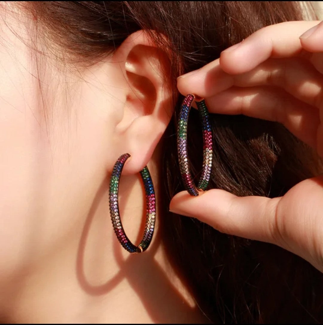 Close-up of a person wearing a large, colorful Shimmer Hoops earring adorned with sparkling zircon stones and holding its matching pair. These affordable luxury hoops dazzle with vibrant colors and eye-catching brilliance.