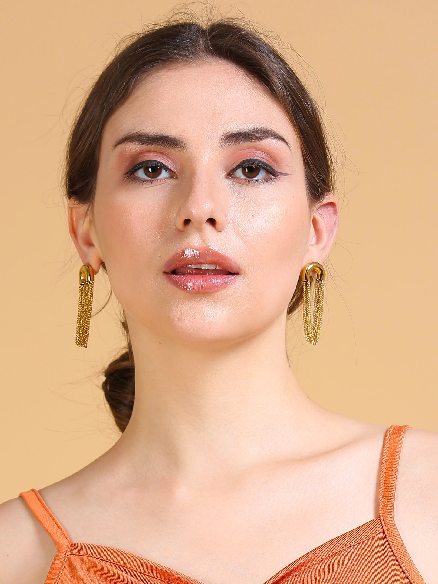 A woman with fair skin and brown hair tied back models Gold chain earrings, a classic jewelry accessory, in front of a plain beige background.