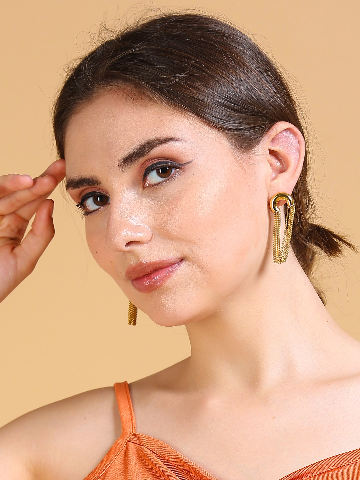 A woman with fair skin and brown hair in a low bun wears gold chain earrings as a statement accessory and an orange sleeveless top, posing against a beige background with one hand raised near her face.
