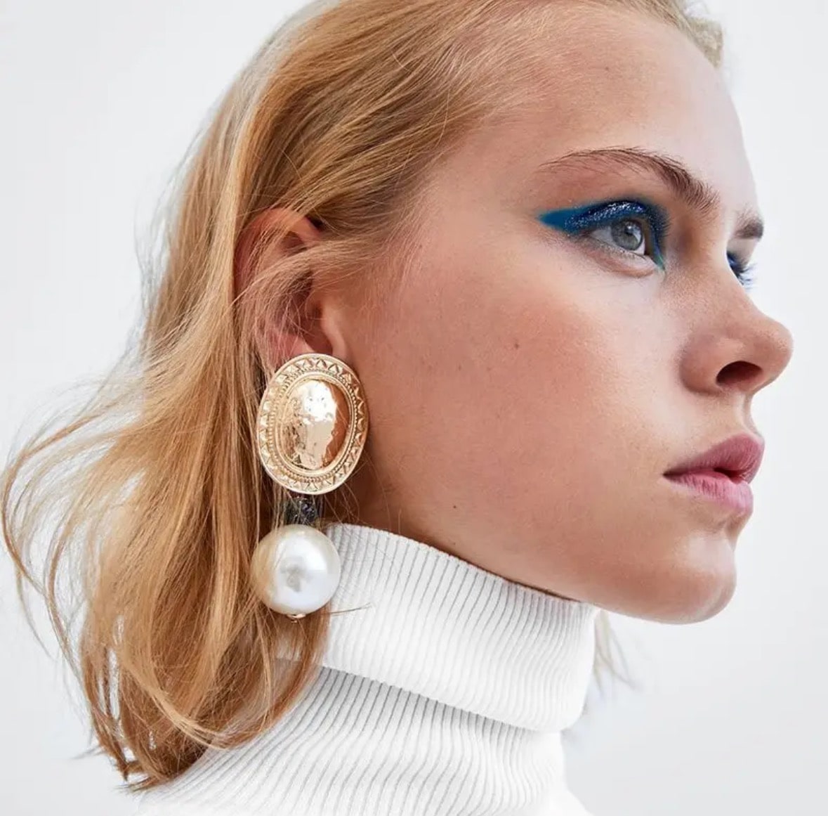 A blonde-haired woman in a white turtleneck, bold blue eyeshadow, and Pearl & Shield Earrings gazes right against a plain light background.