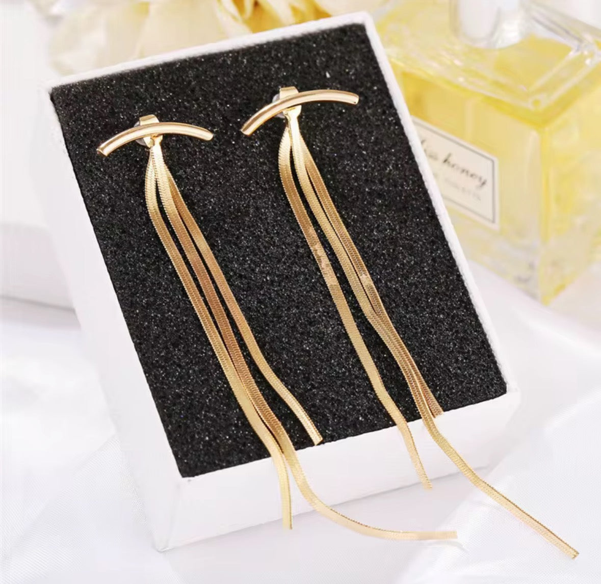 The Tassel no Tassel Earrings are 24K gold plated with long, thin, dangling chains and come in a black velvet jewelry box, placed beside a bottle of perfume.