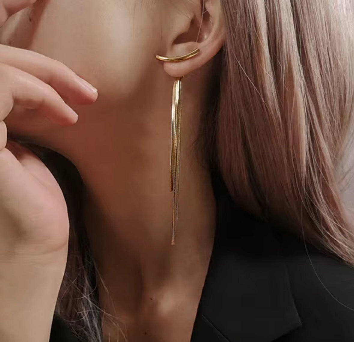 A person with light hair wears Tassel no Tassel Earrings—long, elegant 24K gold plated pieces with thin strands—styled with a black blazer. Their hand is near their face, partially covering their mouth.