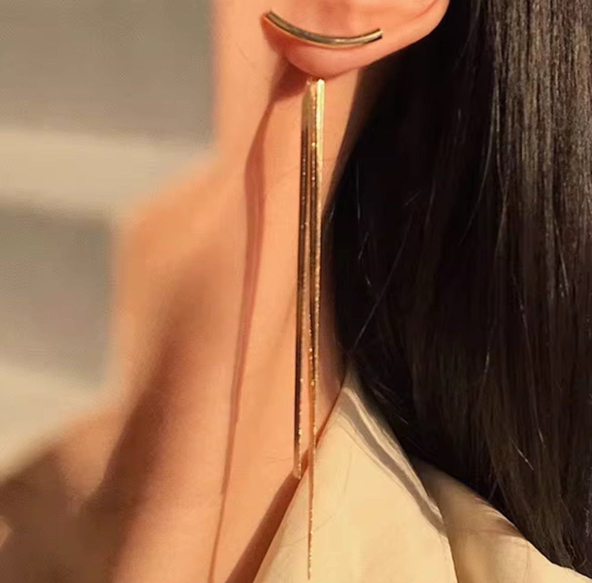 A close-up of a woman's ear wearing Tassel no Tassel Earrings, featuring modern 24K gold plating with a curved bar top and long, thin chains cascading past her shoulder. Her straight dark hair is tucked behind her ear.