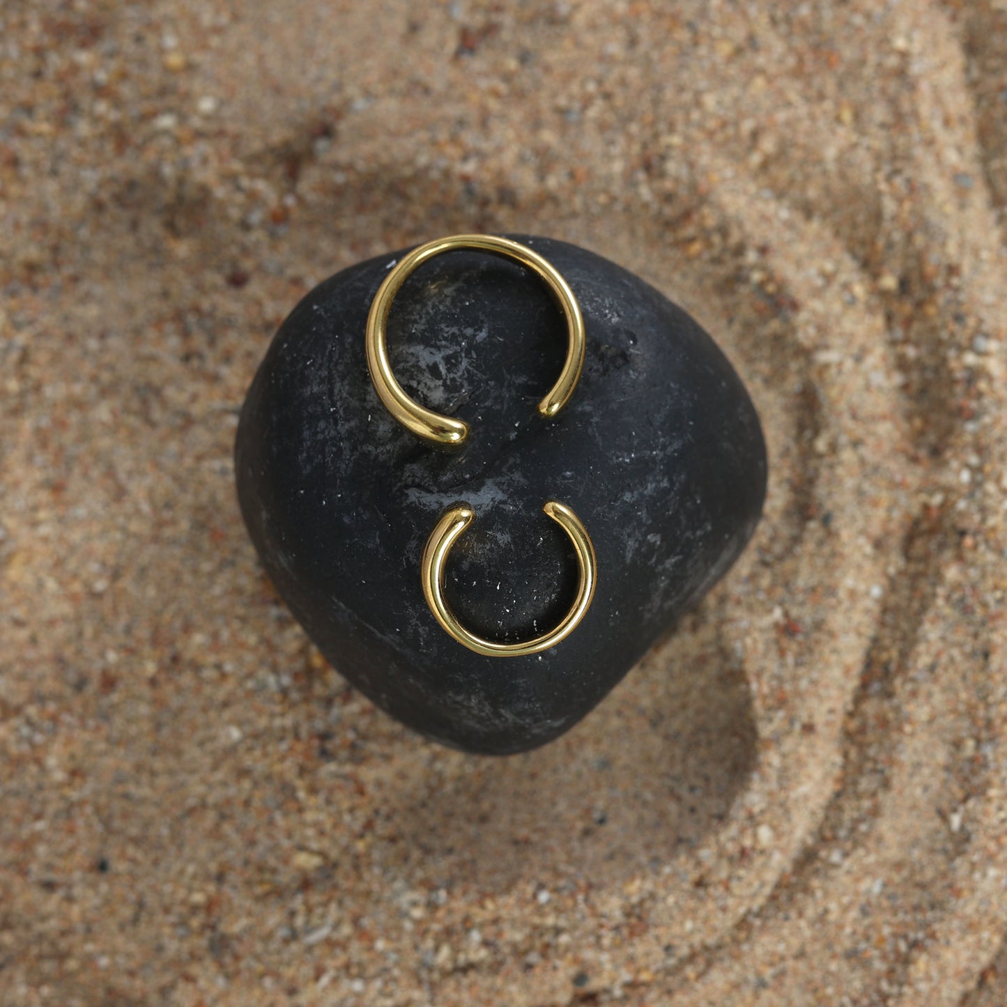 A pair of stylish Gold twin ear cuffs are displayed on a smooth black stone set atop textured sandy ground.