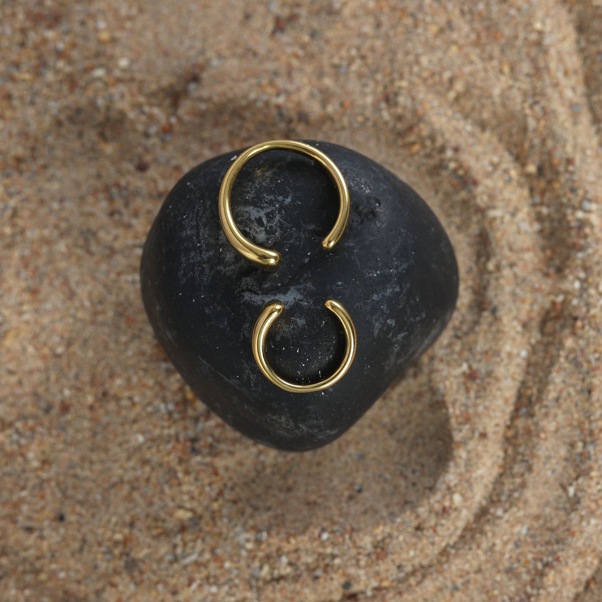 A pair of stylish Gold twin ear cuffs are displayed on a smooth black stone set atop textured sandy ground.