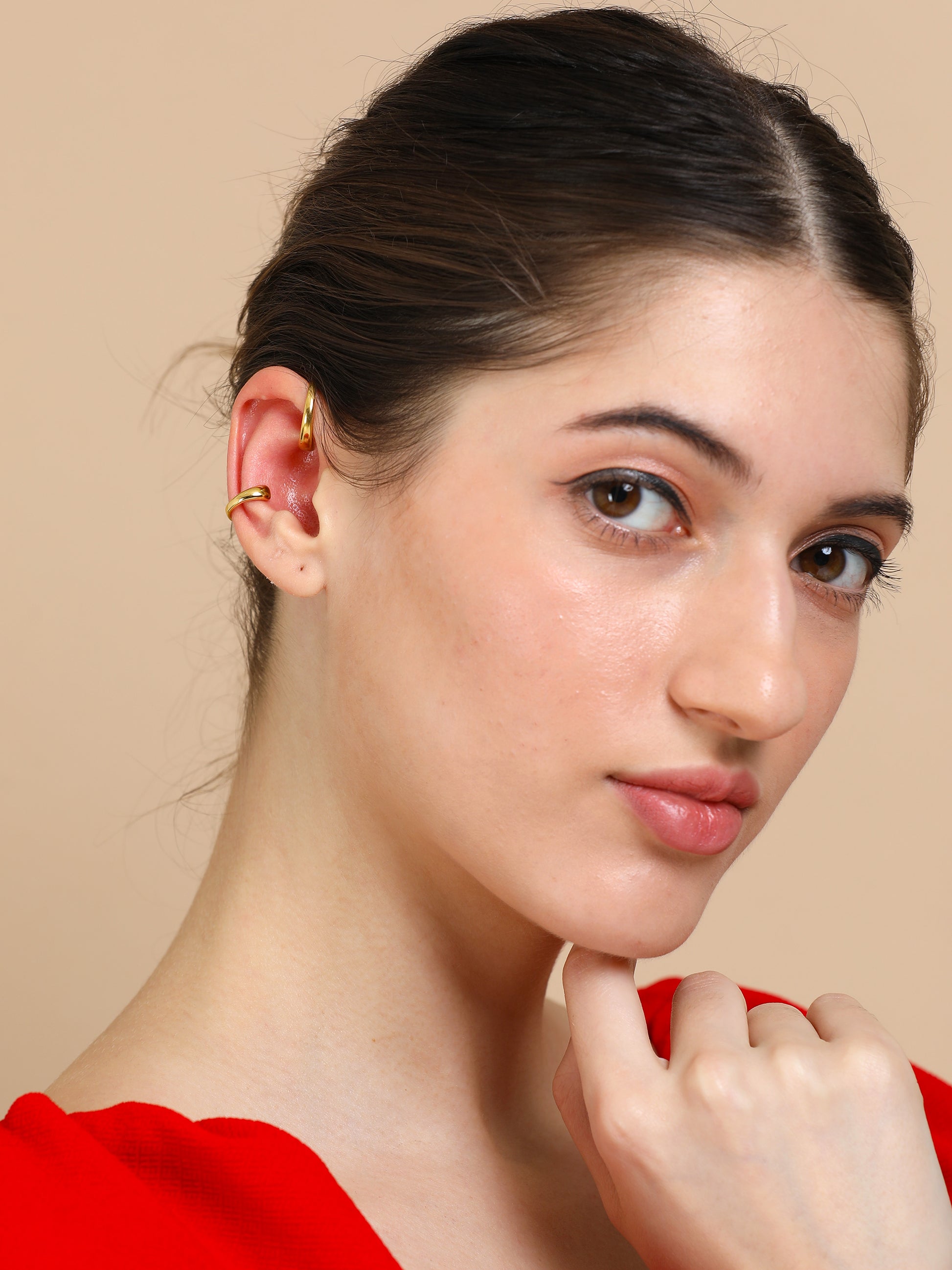 A young woman with light skin and dark hair pulled back, wearing a stylish red top and the Gold twin ear cuff, gazes at the camera with her hand under her chin against a beige background.