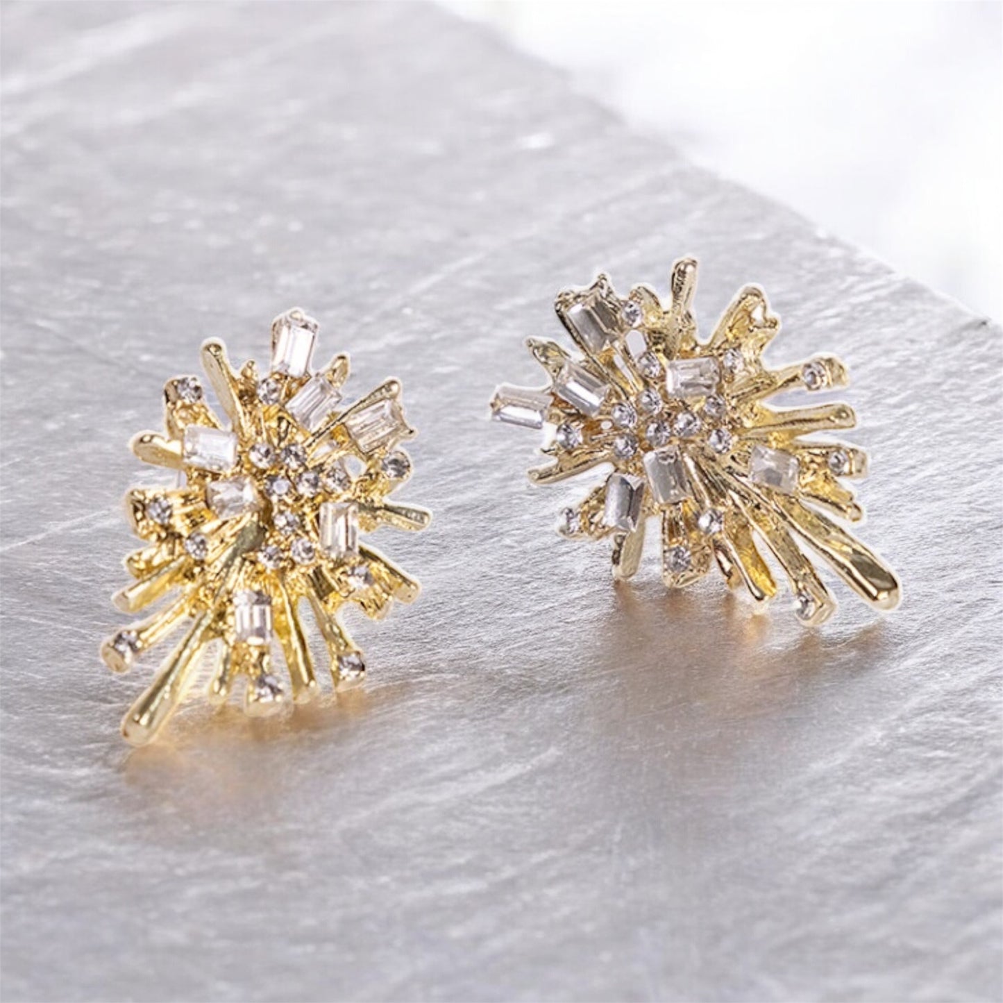 The Gold Firework earrings are 24K gold plated with an abstract starburst design, featuring sparkling lab-created stones and intricate radiating metalwork, displayed on a light textured surface.