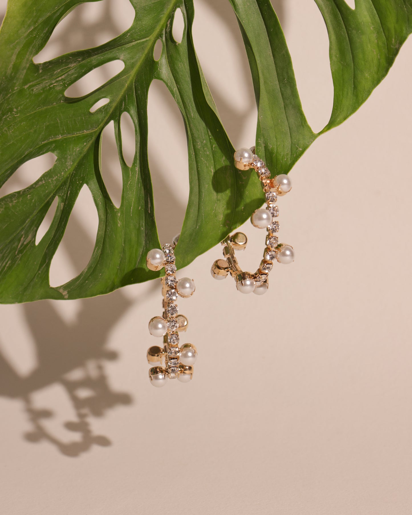 The Gold Pearl and Crystal Hoops, plated in gold and embellished with pearls and crystals, hang from a green monstera leaf, casting a chic shadow on beige—an elegant luxury jewelry accent.